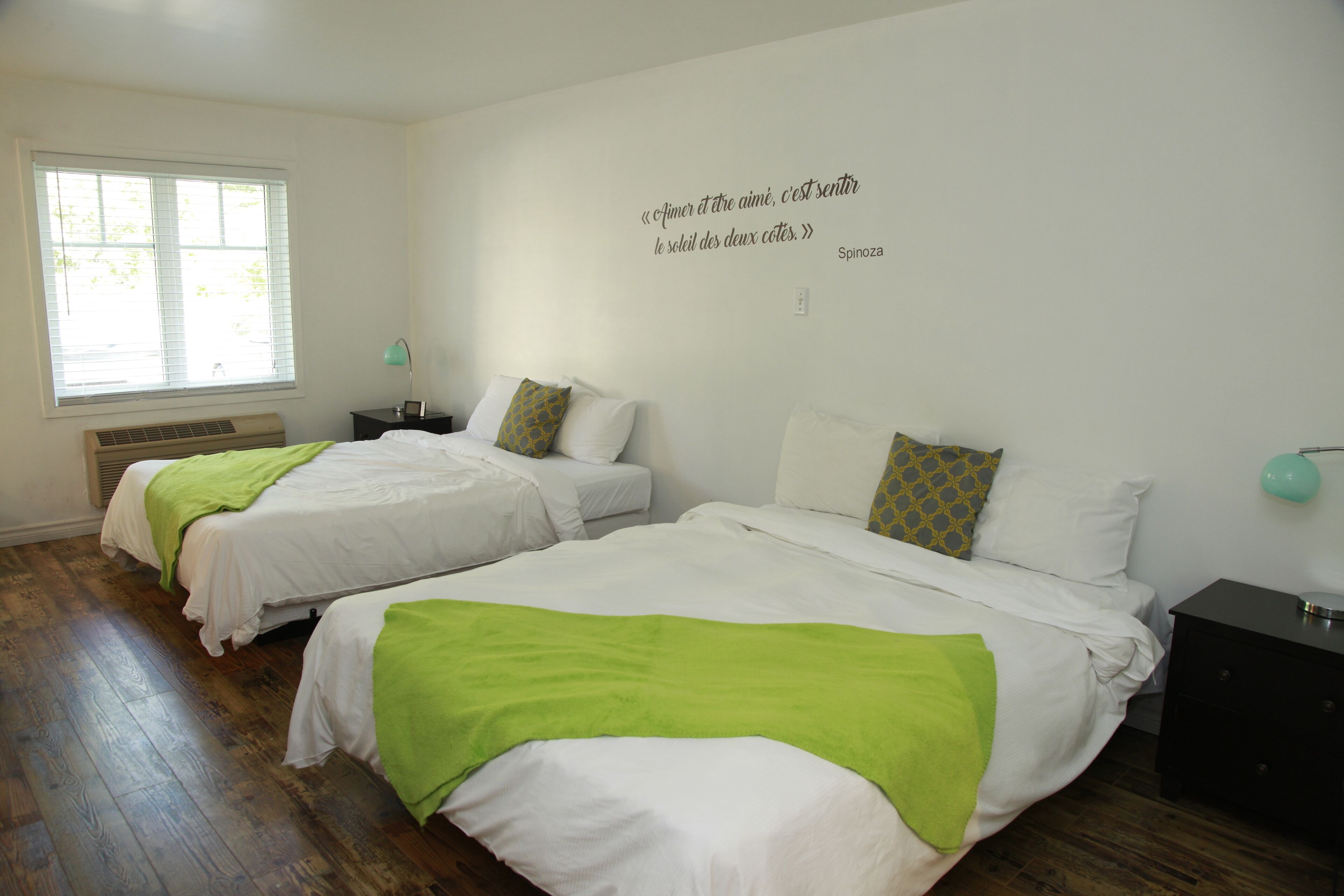 A bedroom with two beds, a window with blinds, a wall quote, and a nightstand with a lamp.