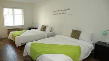 A bedroom with two beds, a window with blinds, a wall quote, and a nightstand with a lamp.