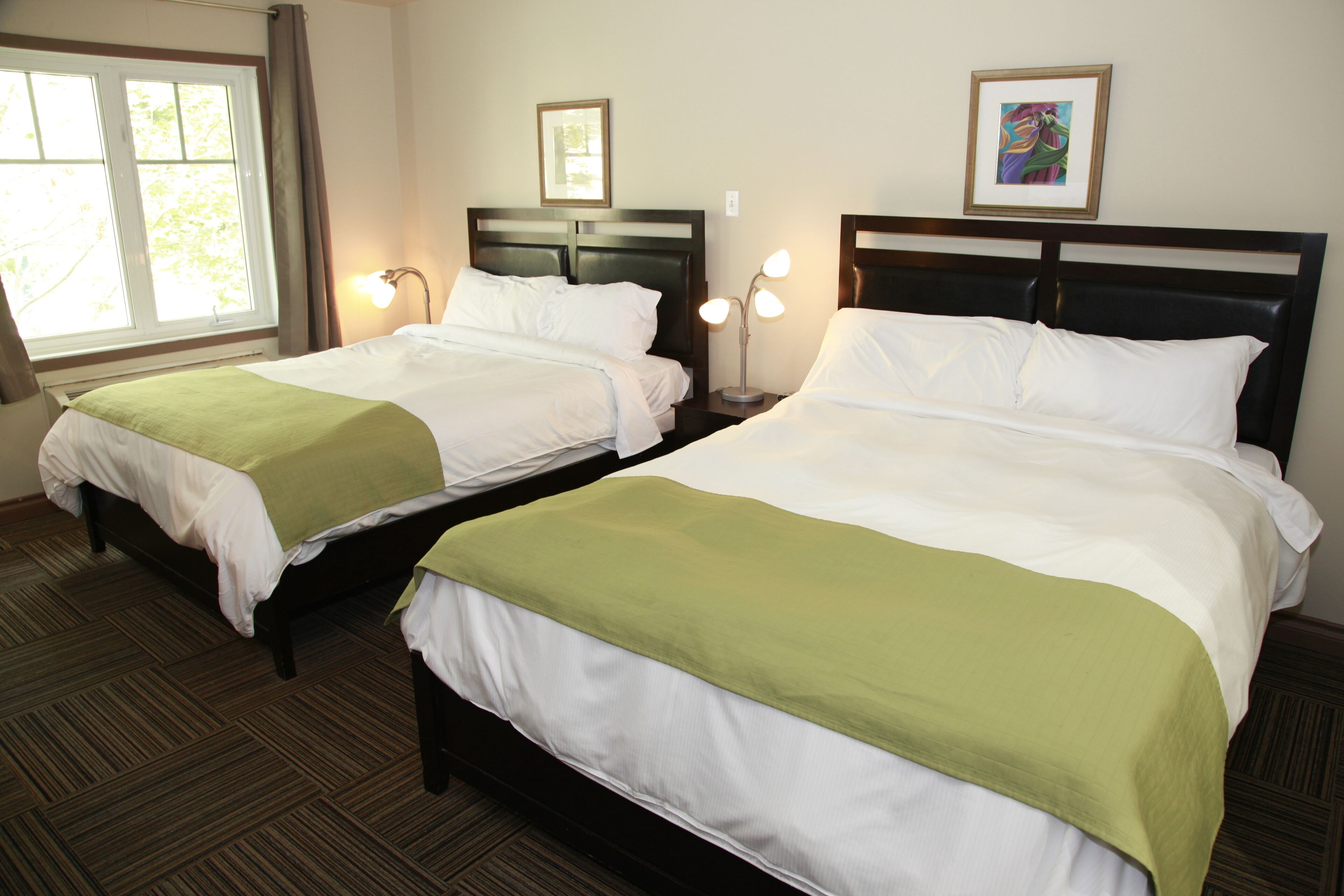 Une chambre d’hôtel avec deux lits doubles, des couvre-lits verts, une grande fenêtre et une table de chevet avec une lampe.