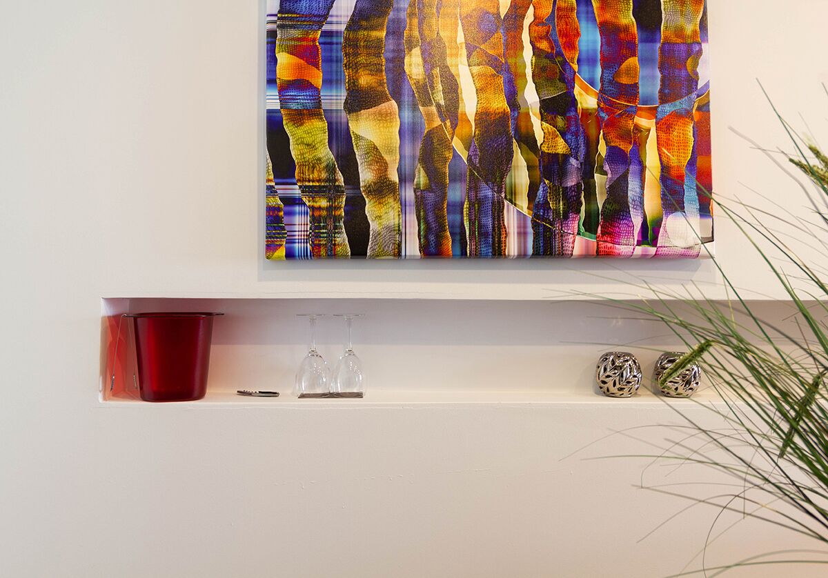 Una pared con un cuadro abstracto colorido, un cubo rojo, dos copas de vino y dos adornos en una repisa.