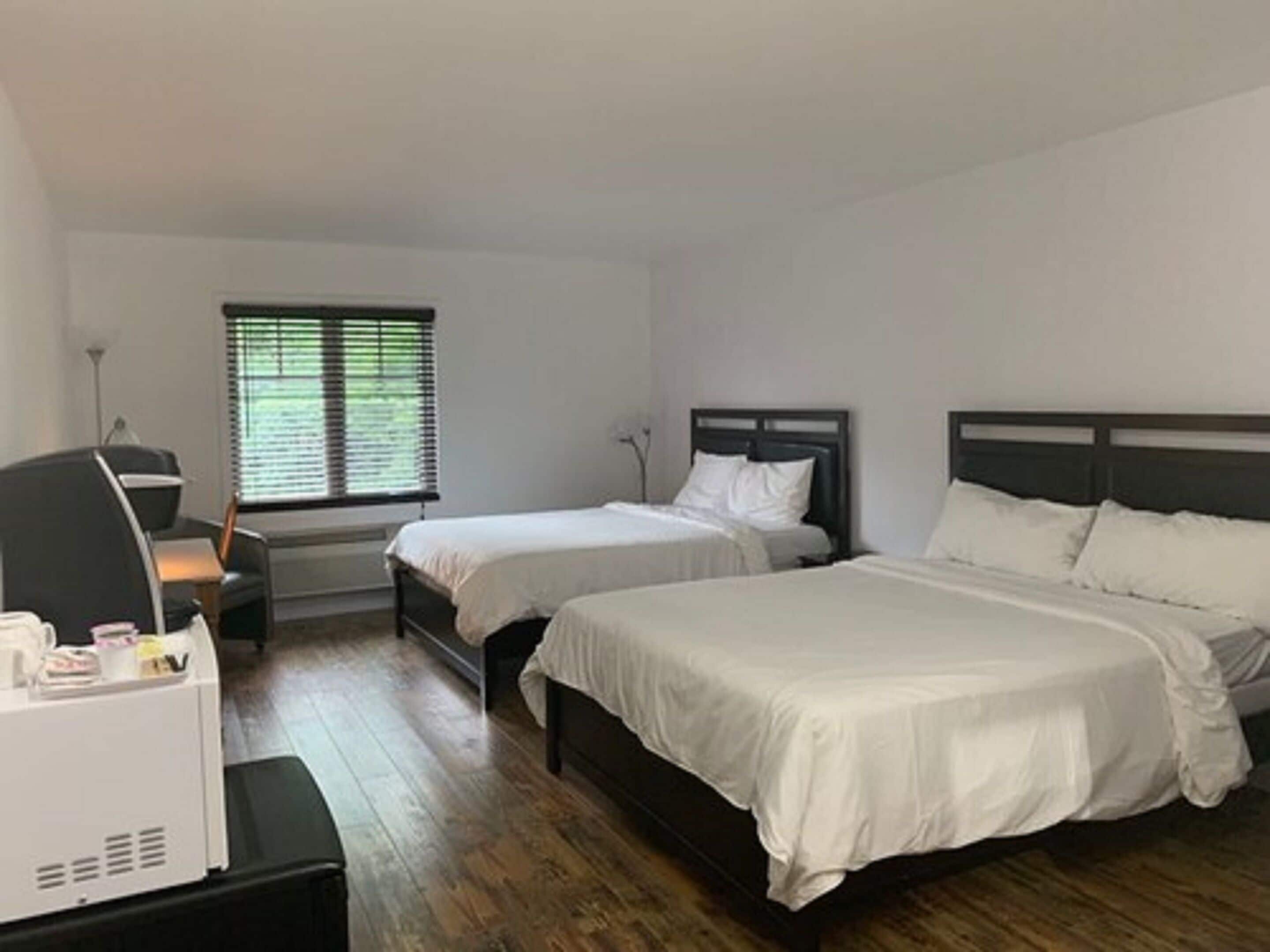 A hotel room with two single beds, a desk with a chair, a window with blinds, and a lamp.
