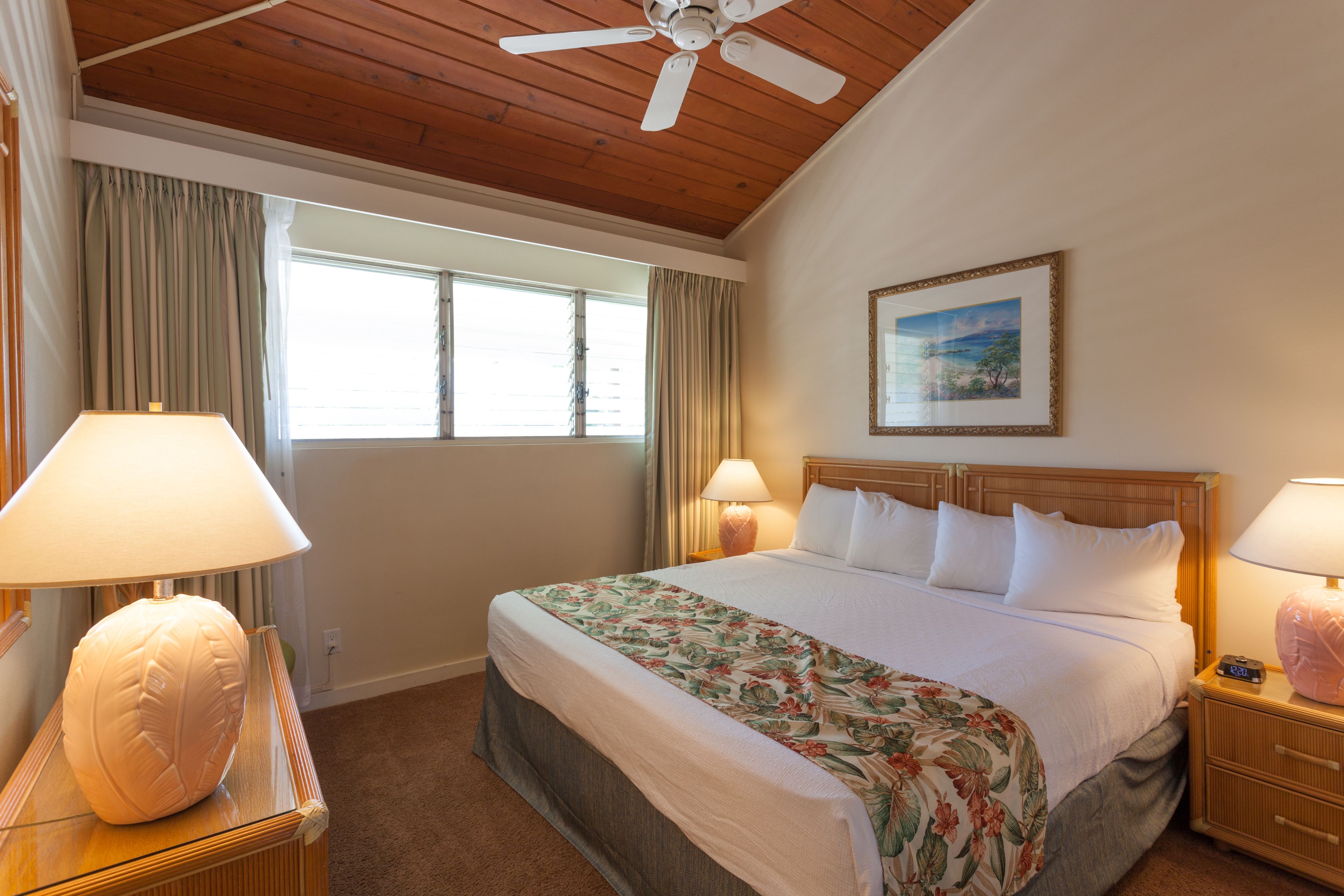 Habitación de hotel con techo de madera, una cama grande con ropa de cama floral, dos lámparas de noche, una mesita de noche con un reloj y una ventana con cortinas.