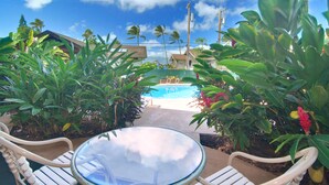 Mesa de vidrio con sillas en un entorno tropical, con vistas a una piscina rodeada de exuberante vegetación.