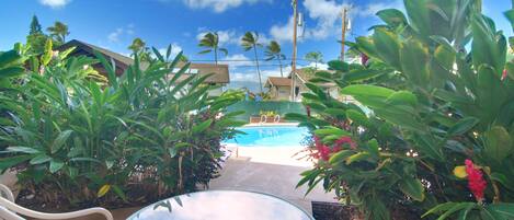 Une table en verre, entourée de chaises, dans un décor tropical, avec vue sur une piscine et bordée d’une végétation luxuriante.