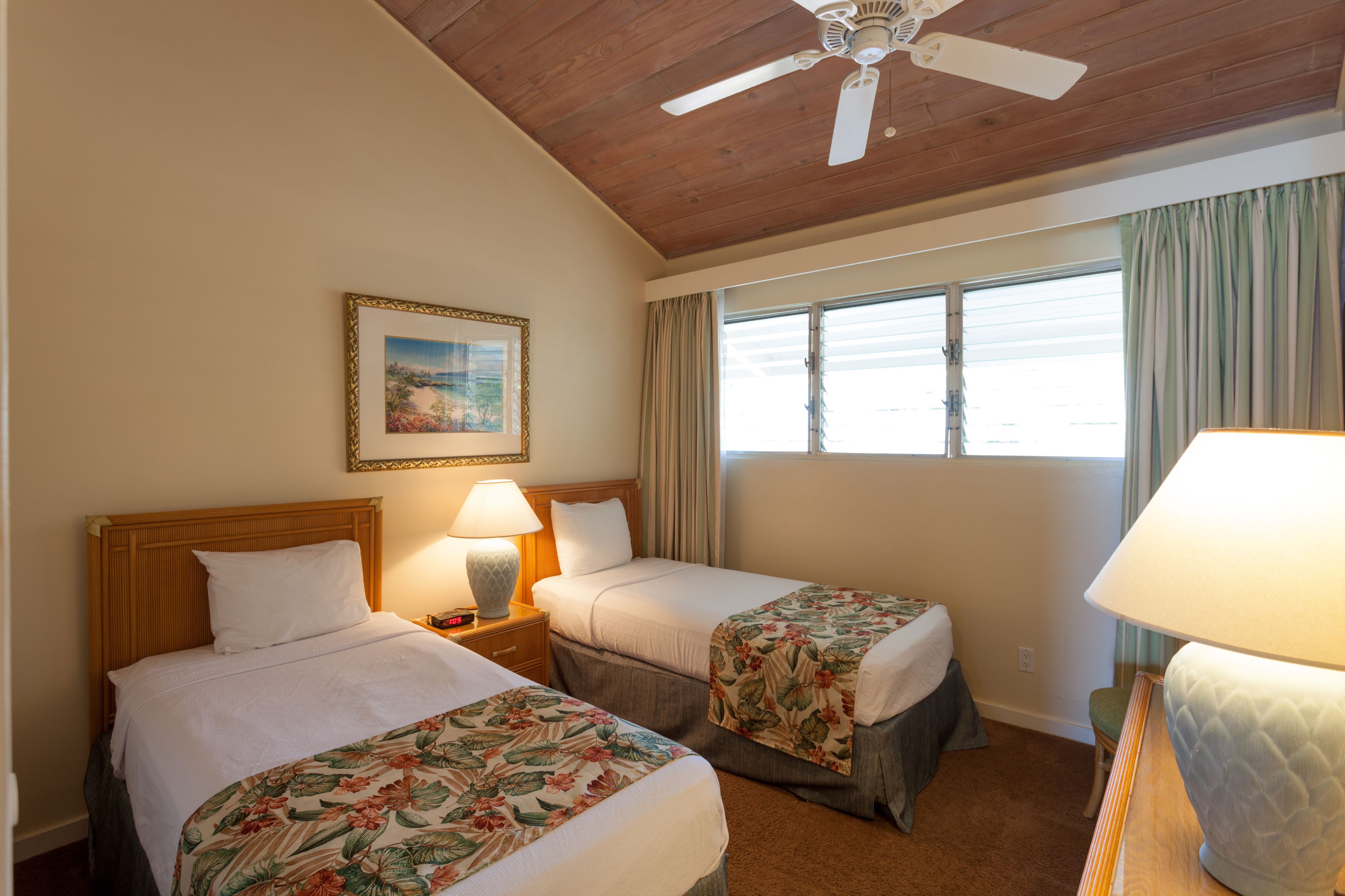 A hotel room with two beds, a ceiling fan, a window with curtains, and a painting on the wall.
