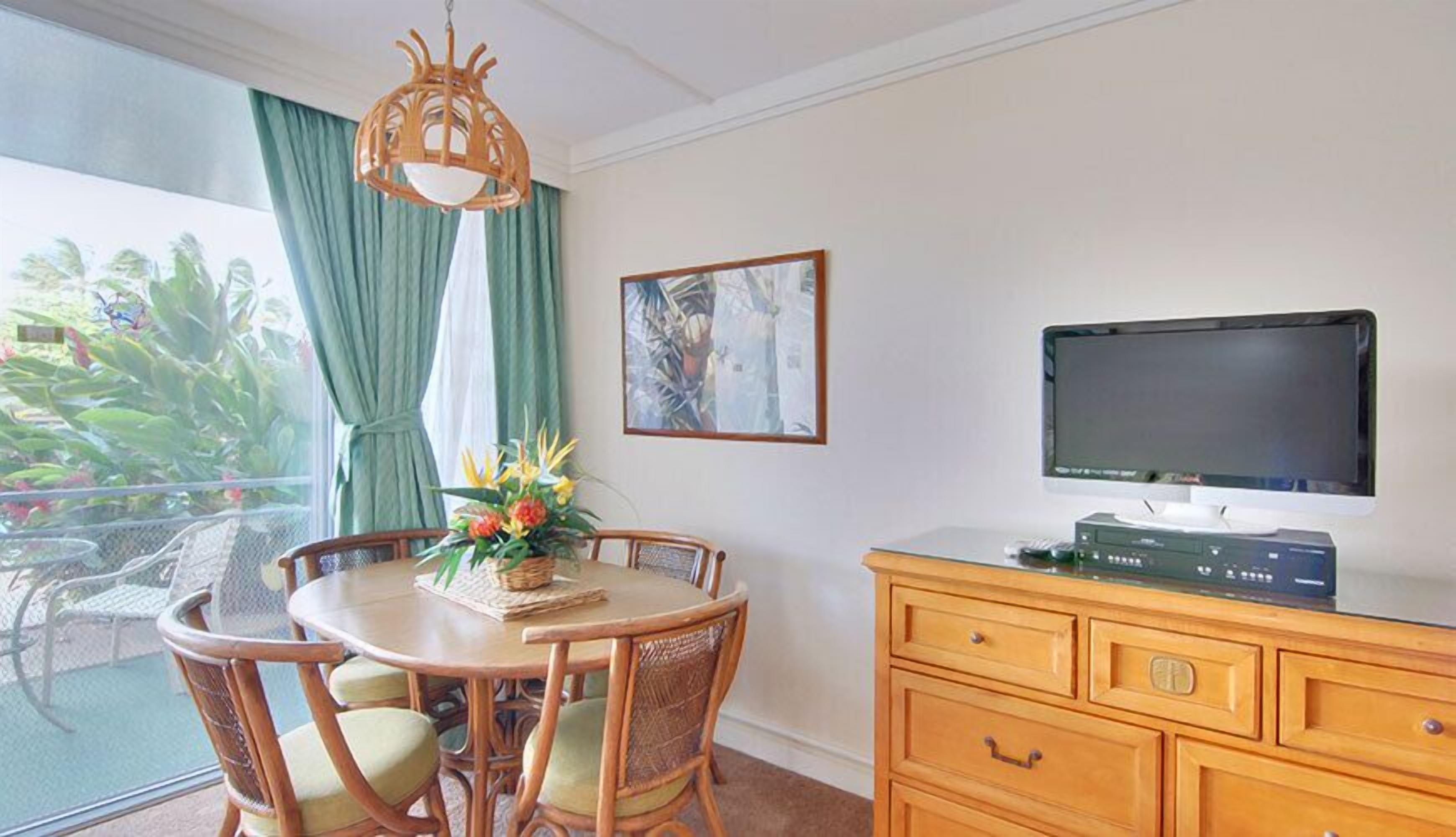 Un coin repas comprenant une table ronde, des chaises, un lustre, un téléviseur posé sur un meuble en bois et une fenêtre avec des rideaux.