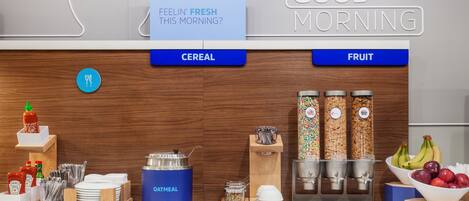Una estación de desayuno con opciones de cereales y fruta, un dispensador de kétchup y un cartel que dice “GOOD MORNING”.