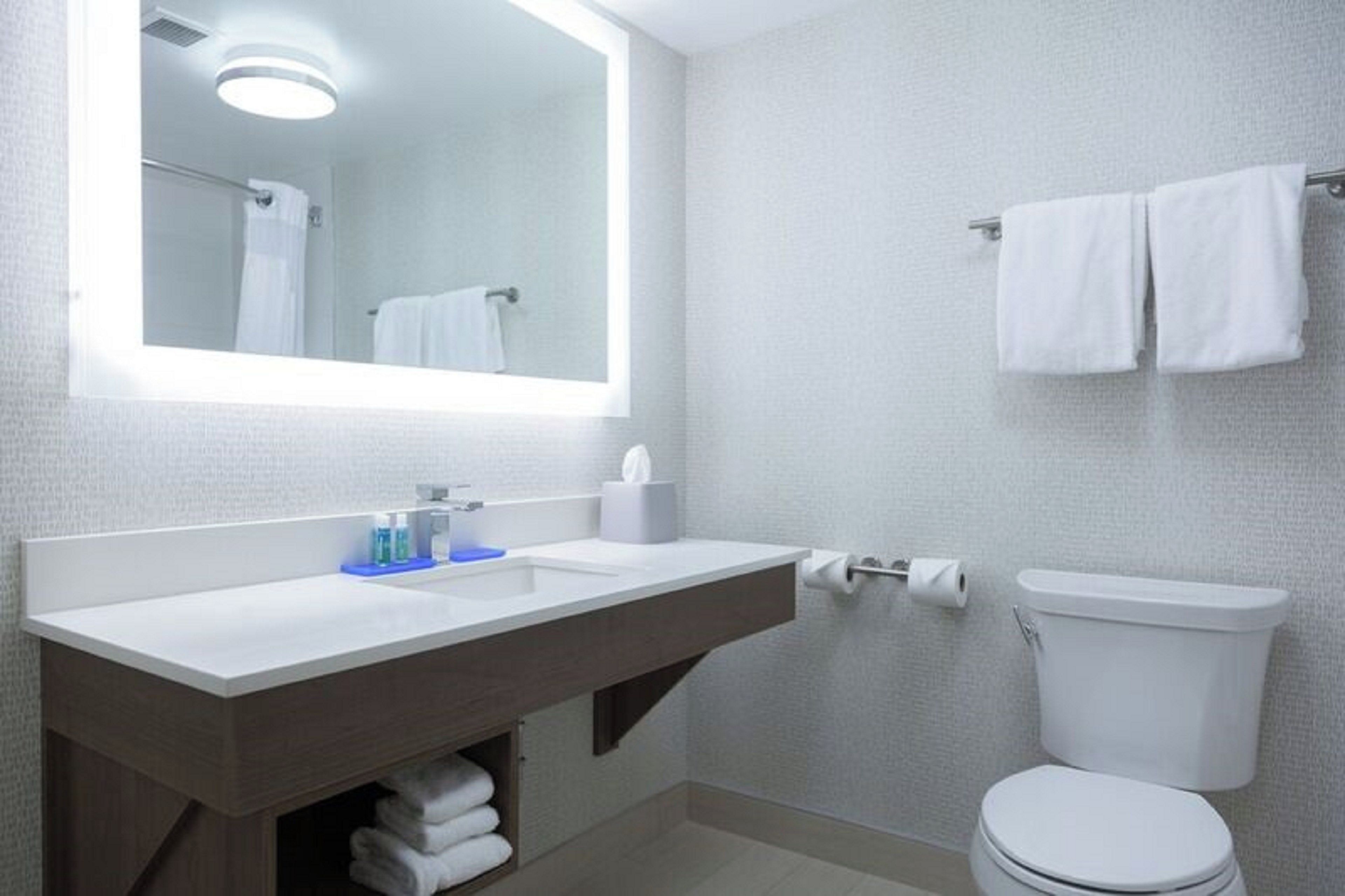 A bathroom with a white sink, mirror, and toilet. There are towels hanging on the wall and a shelf with toiletries.