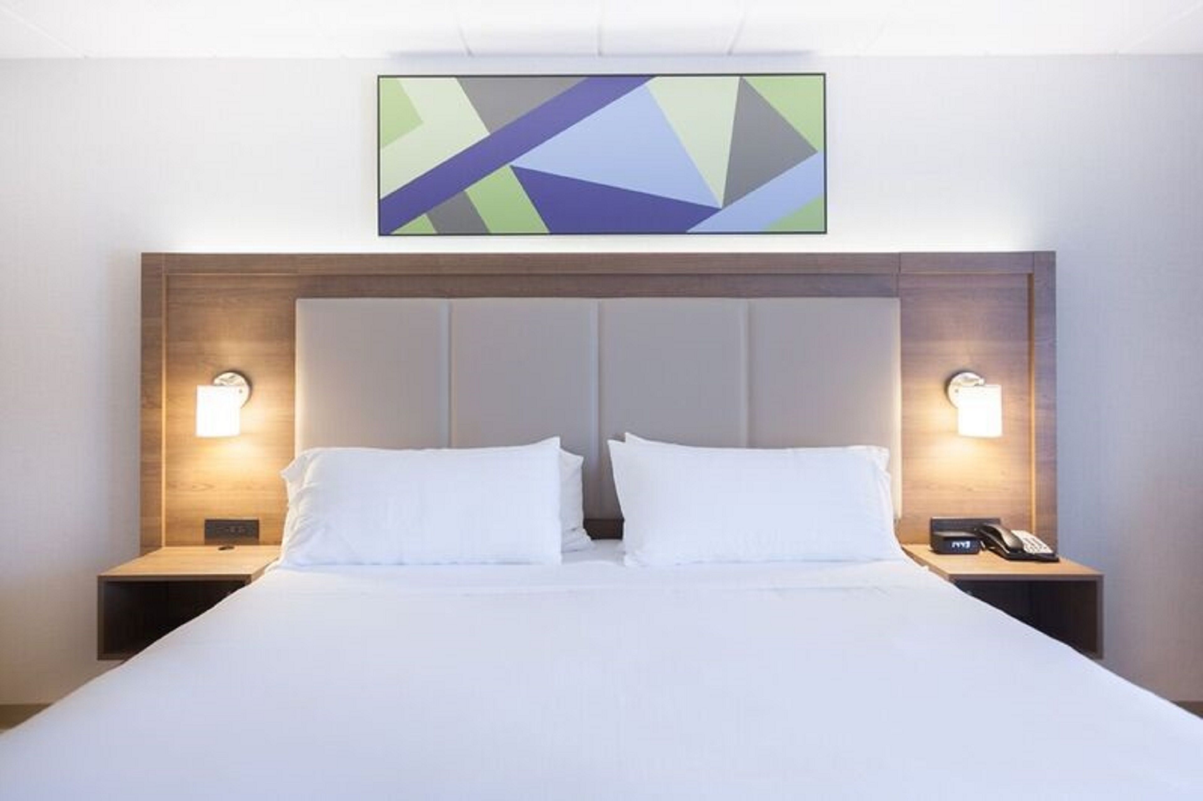 Habitación de hotel con una cama grande, dos mesitas de noche con lámparas, un cabecero moderno y una obra de arte geométrica en la pared.