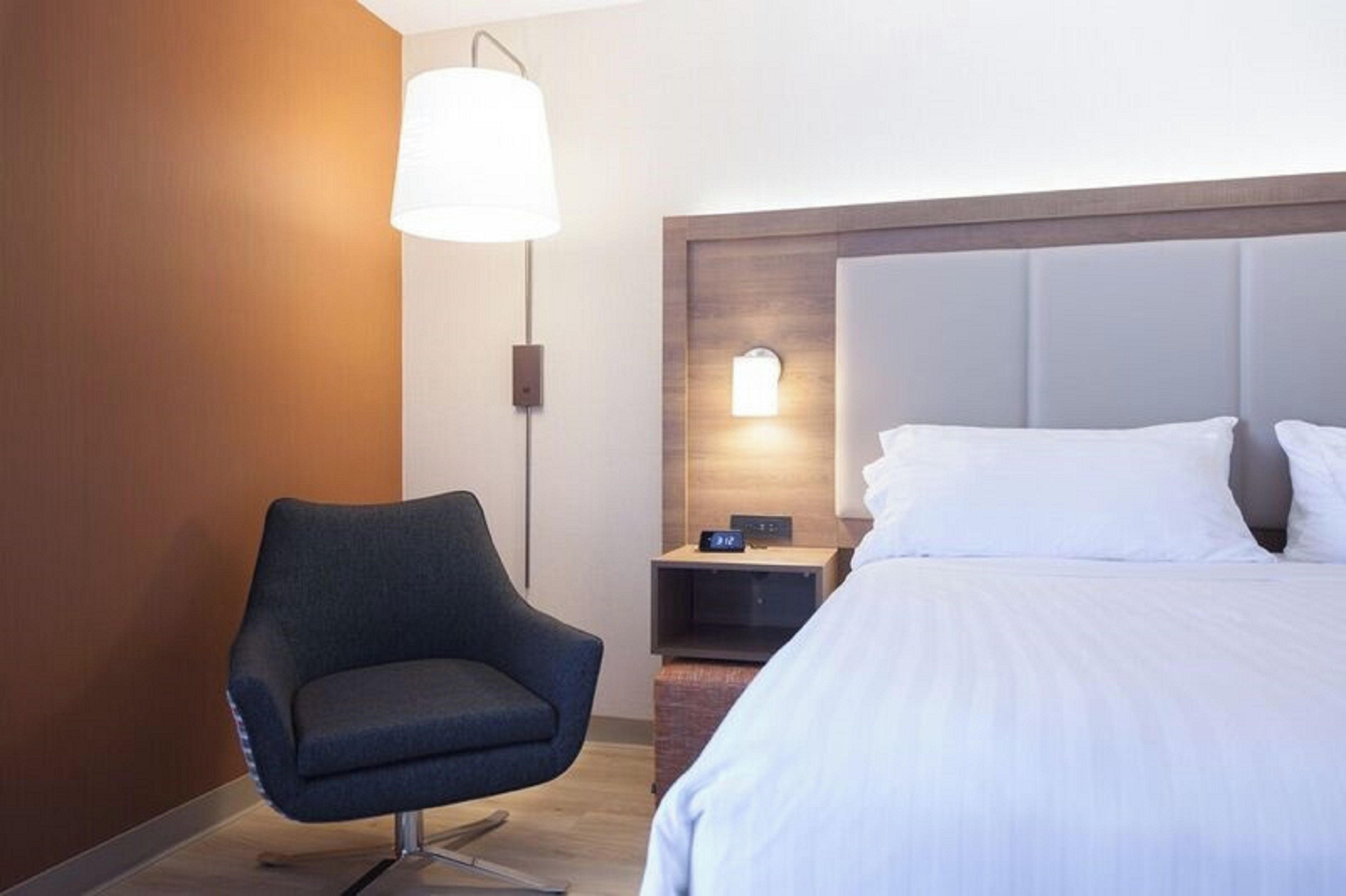 Une chambre d’hôtel avec un lit, une chaise, une table de chevet et une lampe fixée au mur.