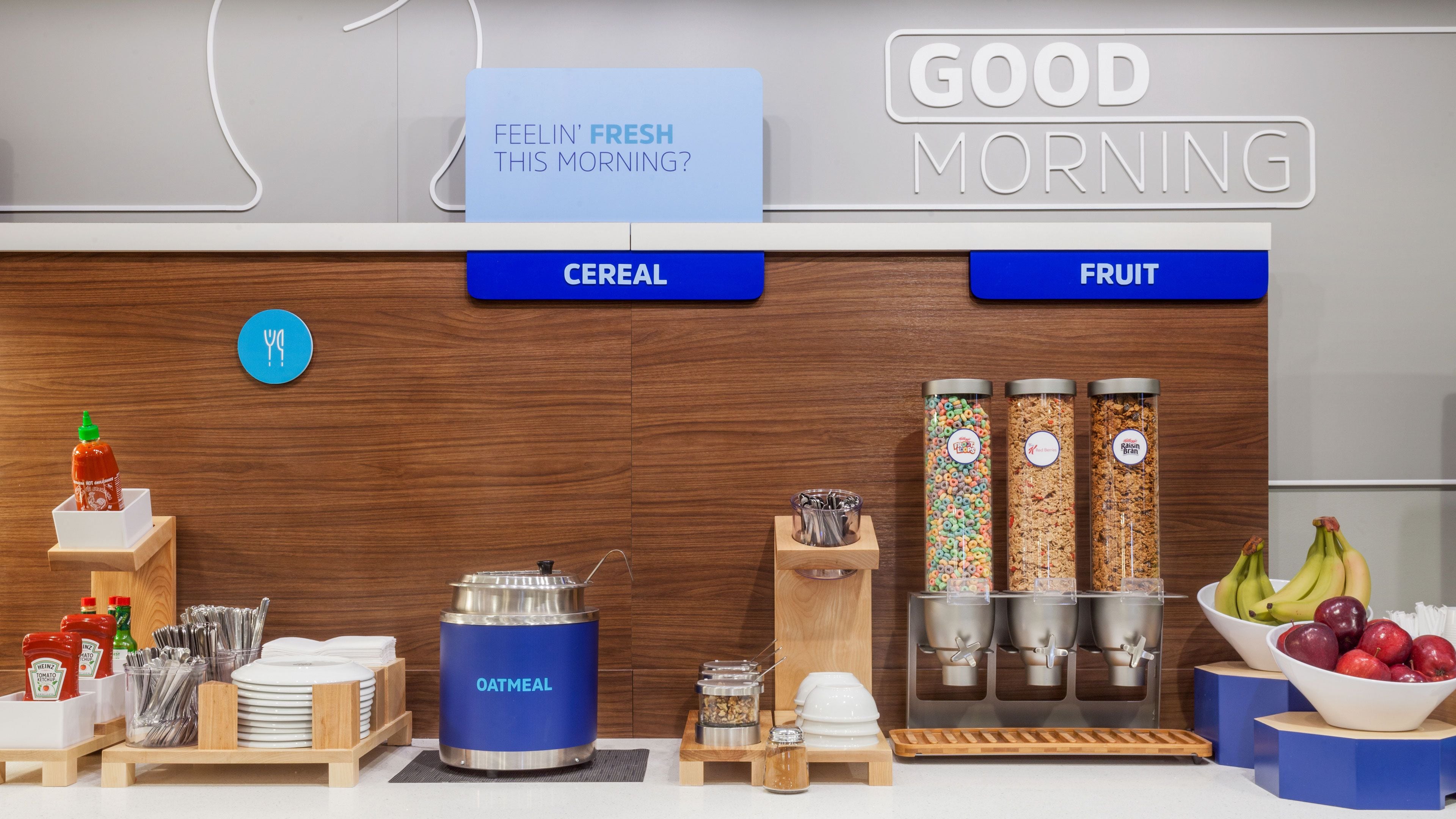 Una barra de desayuno con cereales y frutas, un dispensador de kétchup y un cartel que dice 'GOOD MORNING'.