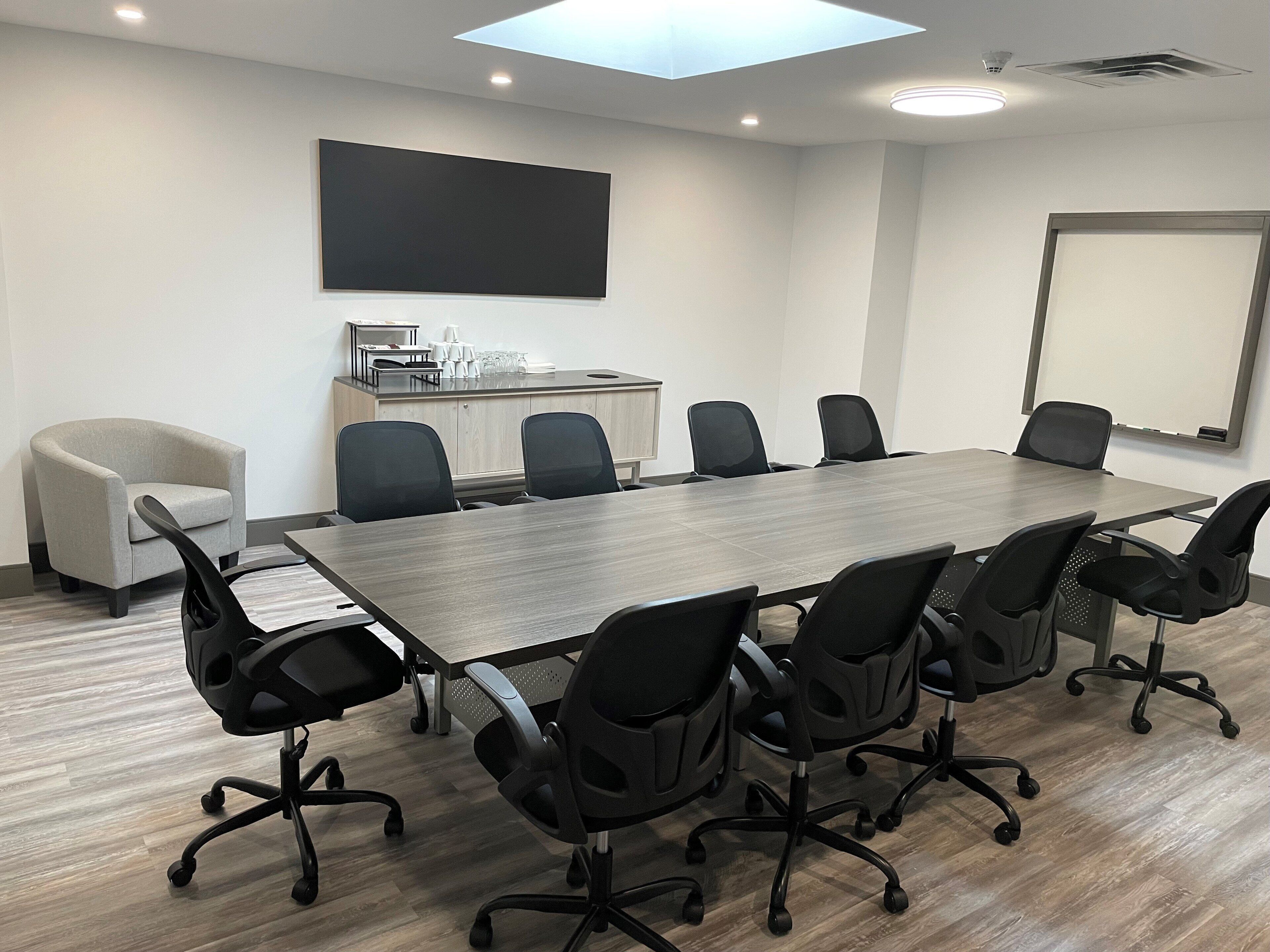 Uma sala de conferências com uma mesa comprida, cadeiras de escritório pretas, um pequeno sofá, um quadro branco e uma TV de tela plana na parede.
