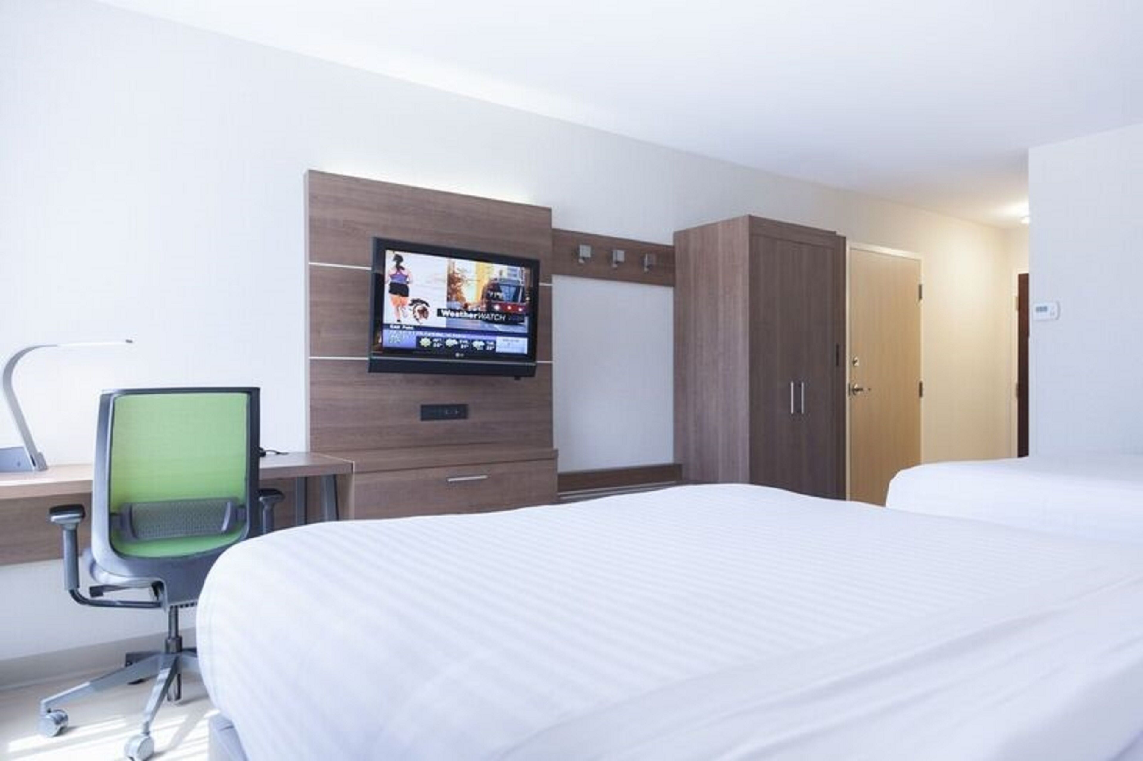Una habitación de hotel con una cama, un escritorio con silla, un televisor montado en la pared y un armario.