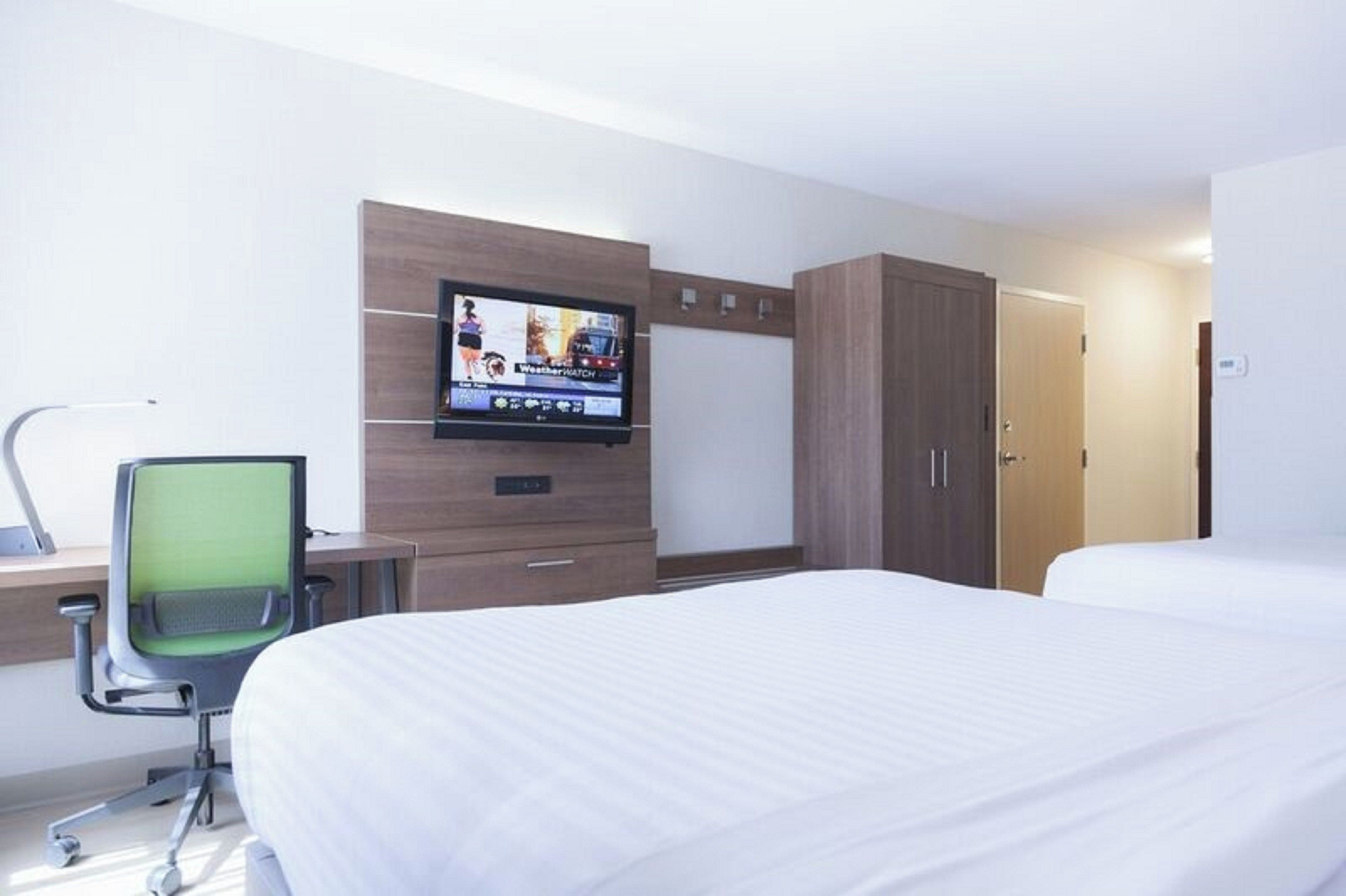 A hotel room with a bed, a desk with a chair, a television mounted on the wall, and a wardrobe.