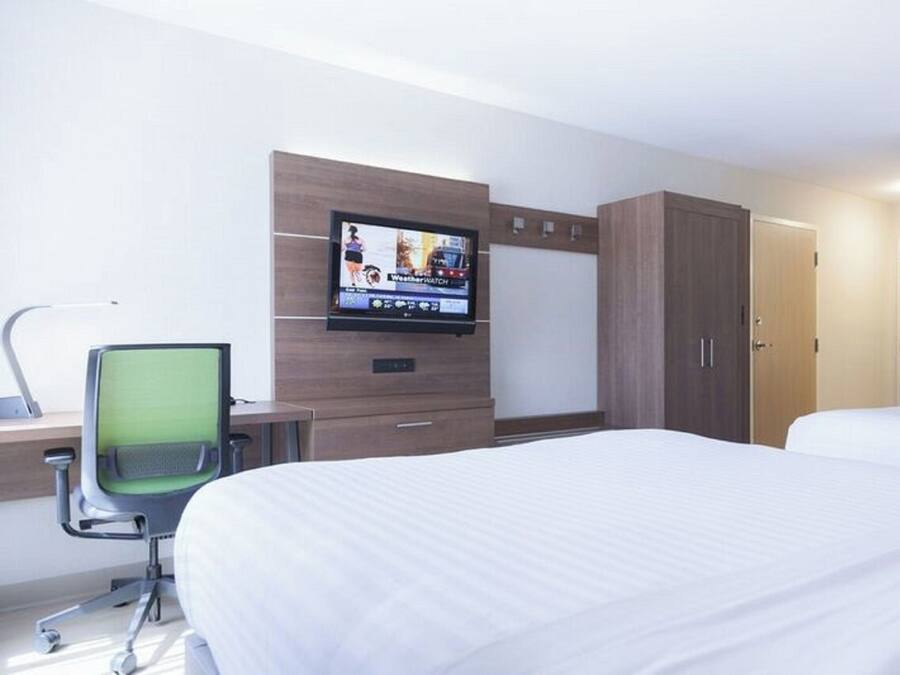 A hotel room with a bed, a desk with a chair, a television mounted on the wall, and a wardrobe.