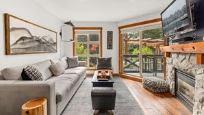 Una sala de estar moderna con un sofá, un reposapiés y una chimenea. Hay un televisor montado en la pared y una ventana con vistas al exterior.