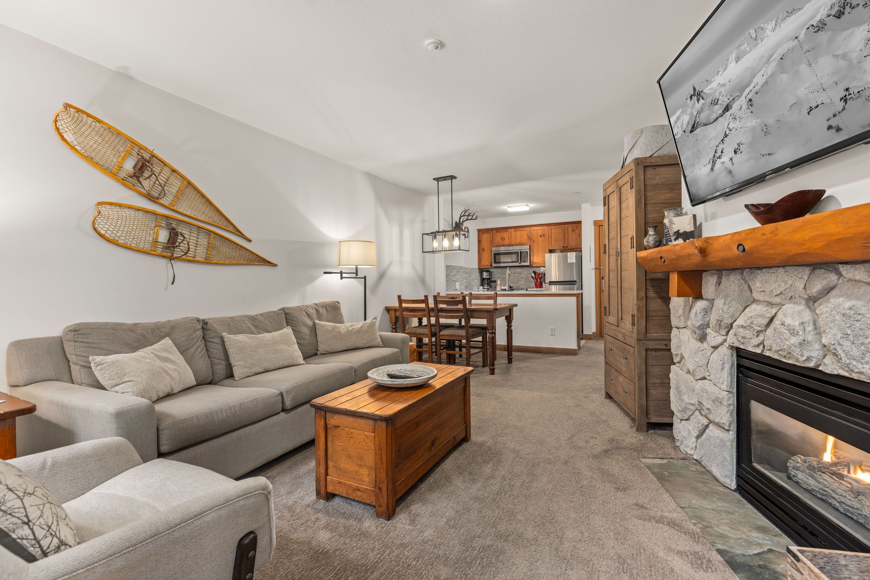 A living room with a sofa, coffee table, and fireplace. There is a kitchen in the background with wooden cabinets and a stone fireplace.