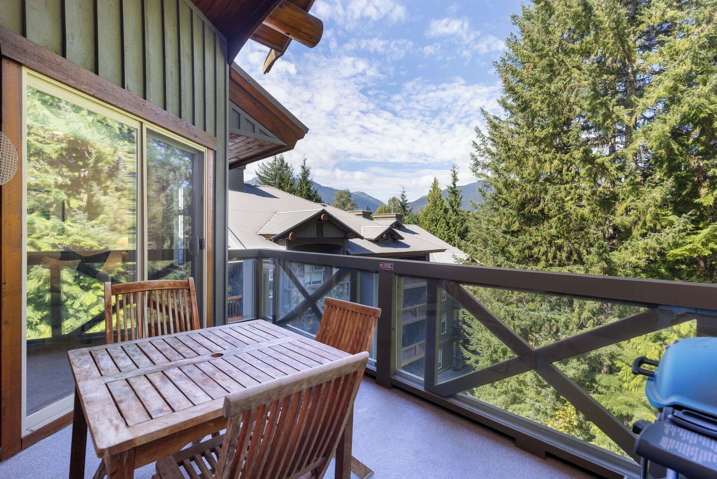 A wooden deck with a table and chairs, overlooking a forested area and mountains.