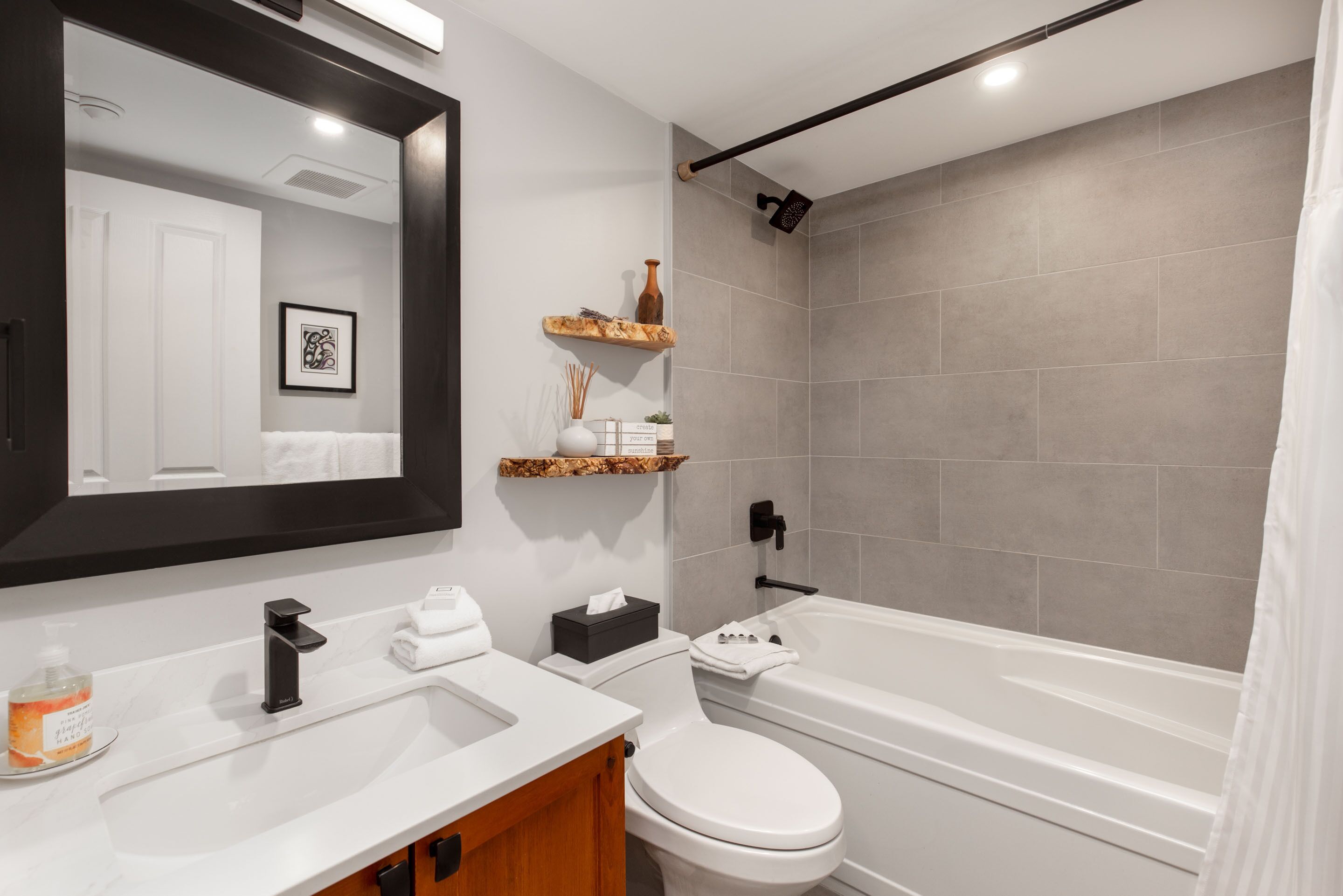 A modern bathroom with a white sink, black faucet, white toilet, and a bathtub. There are wooden shelves with decorative items and a framed picture on the wall.