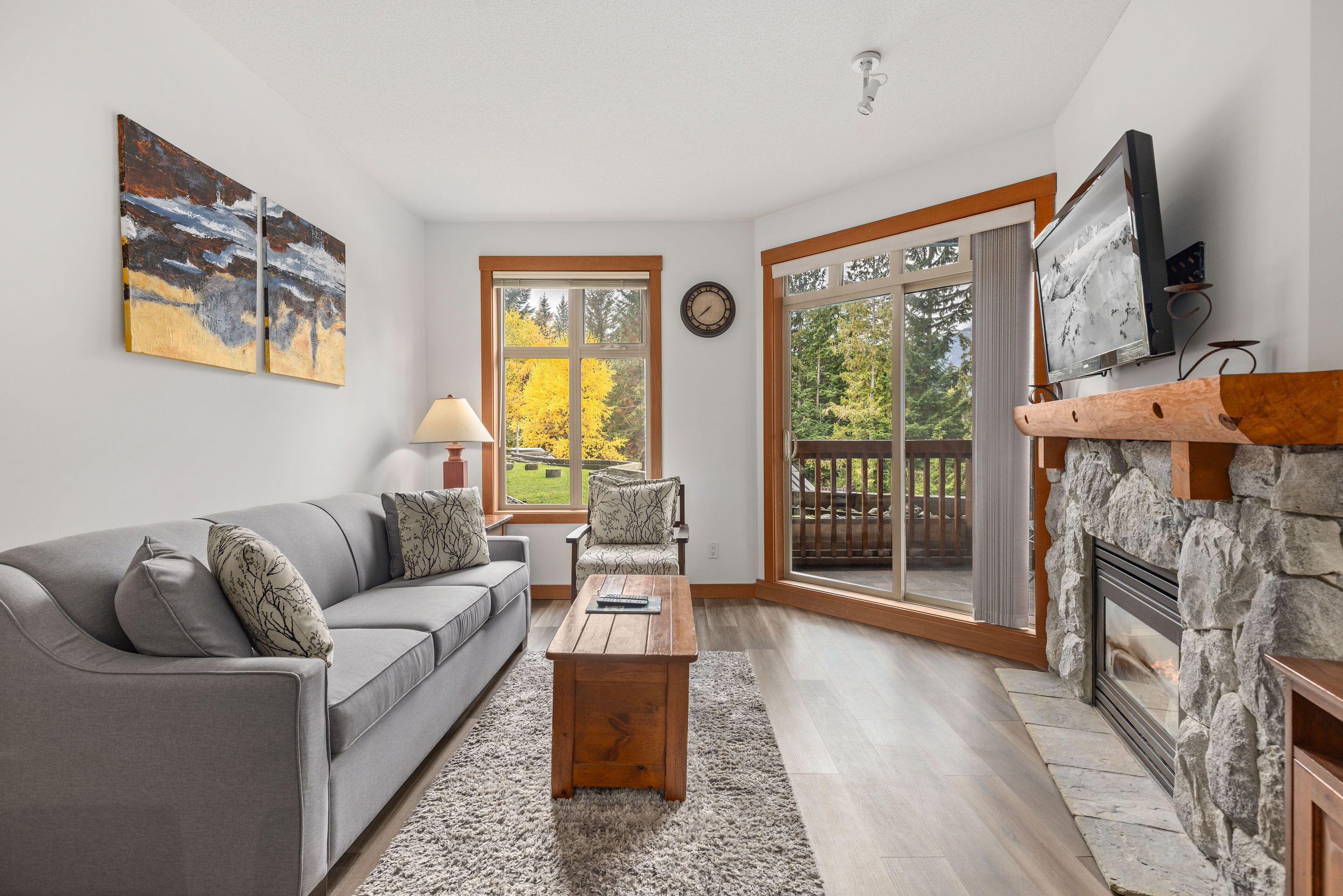 A living room with a gray sofa, a wooden coffee table, a stone fireplace, and a large window with a view of trees.