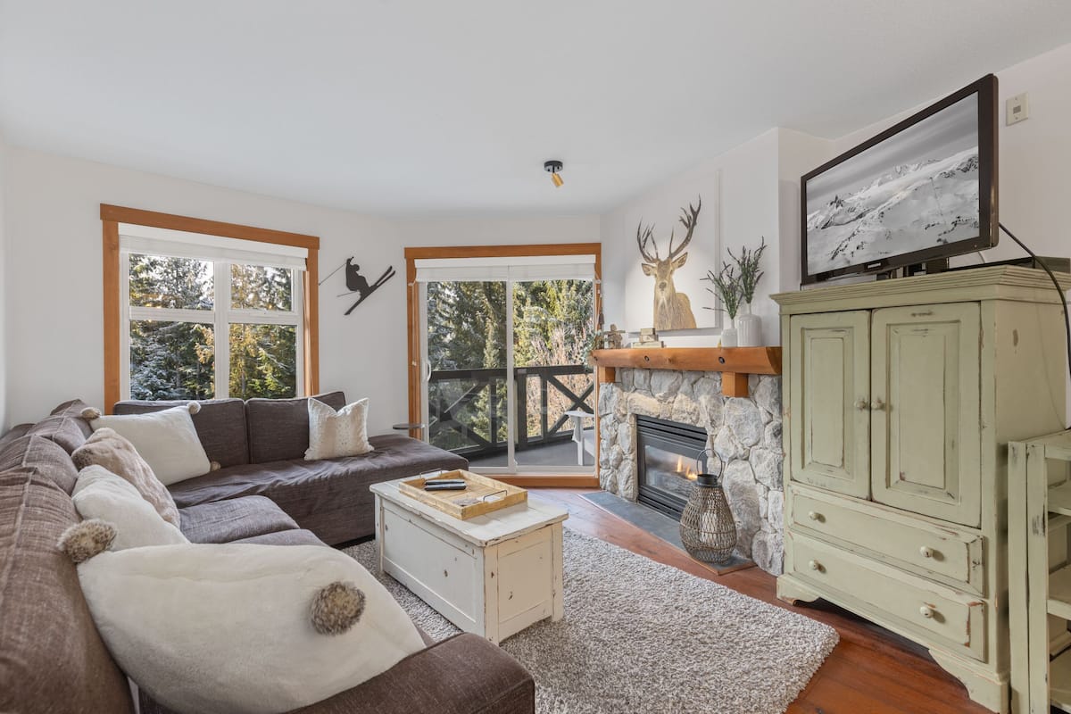 Una sala de estar acogedora con un sofá modular, una chimenea, un televisor y vistas al exterior.