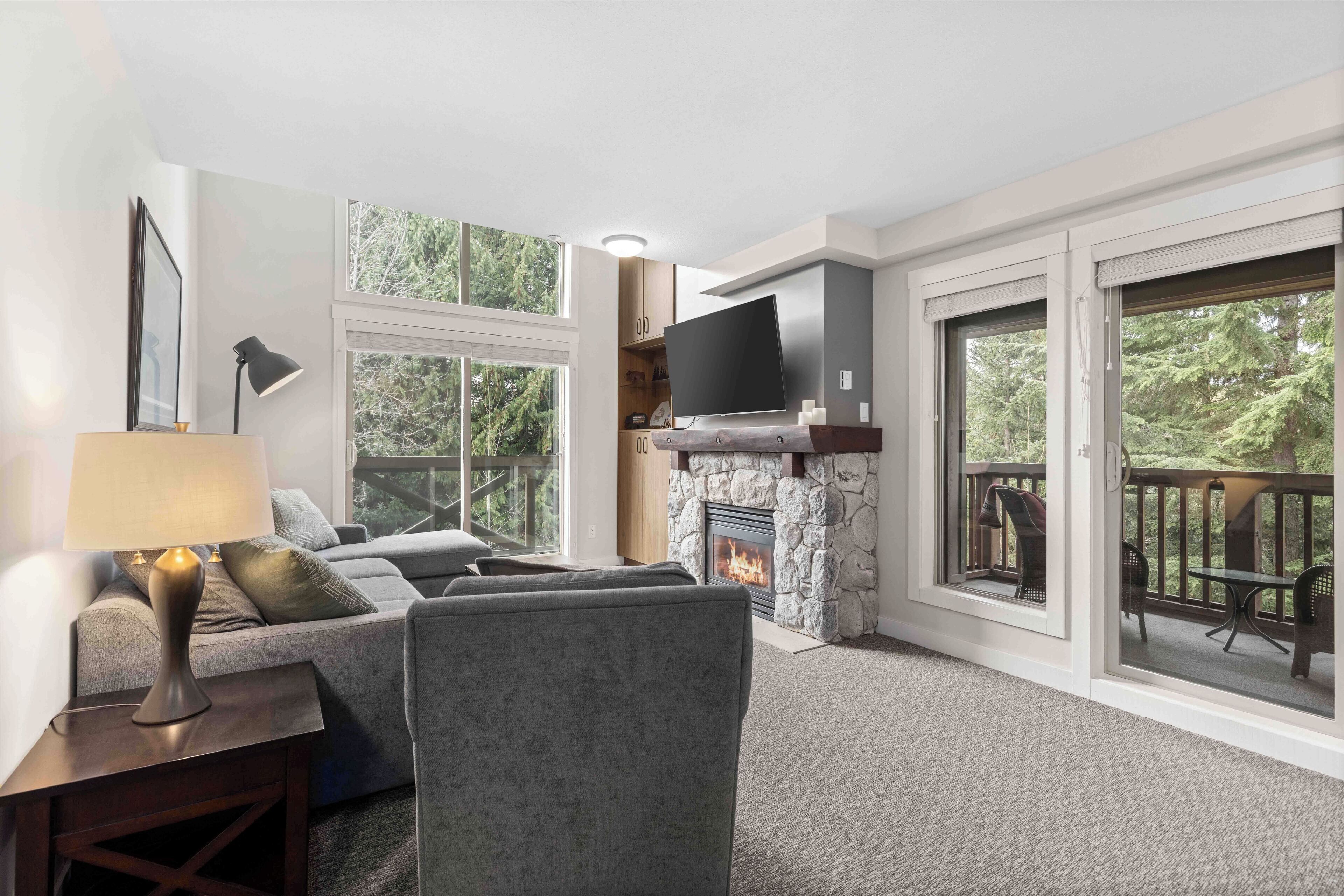 A living room with a stone fireplace, a flat-screen TV mounted above it, and a balcony with outdoor furniture.