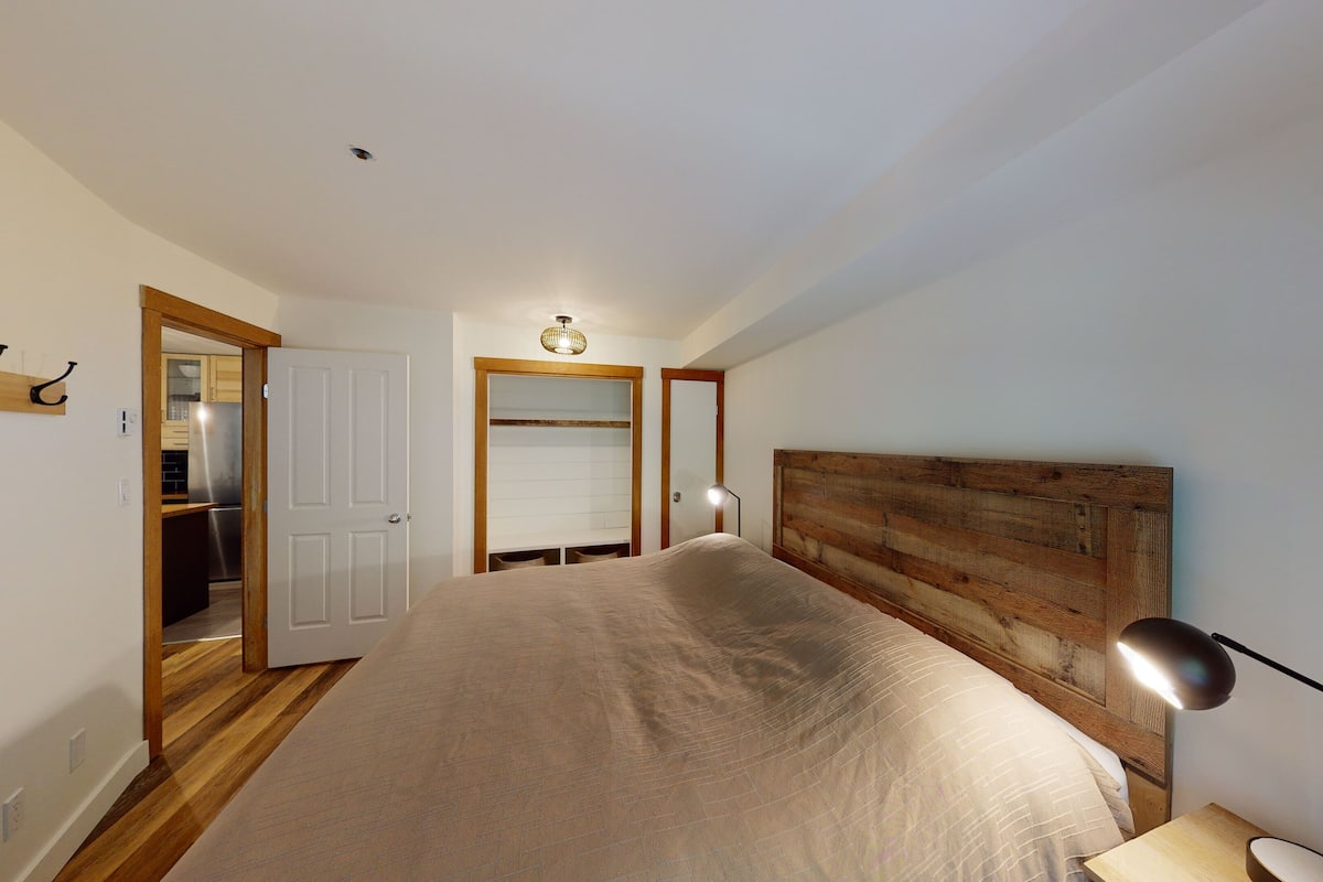 Un dormitorio con cabecera de madera, cama con manta color beige, lámpara de noche y puerta que da al baño.
