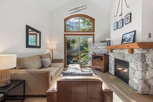A cozy living room with a stone fireplace, a large window with a view of trees, a sofa, and a coffee table.