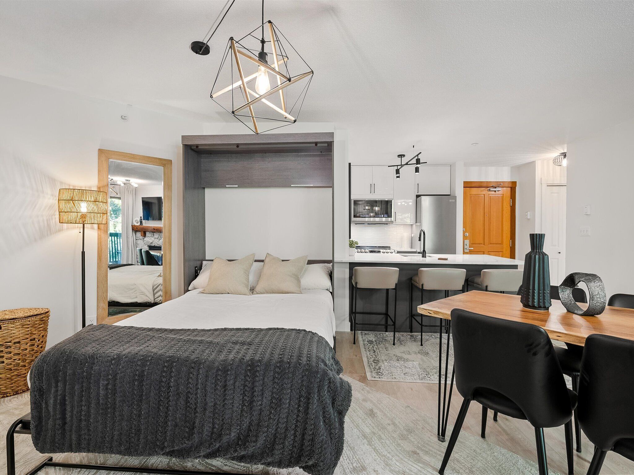 A modern, compact bedroom with a large bed, a wooden dining table, and a kitchen area with a microwave and refrigerator.