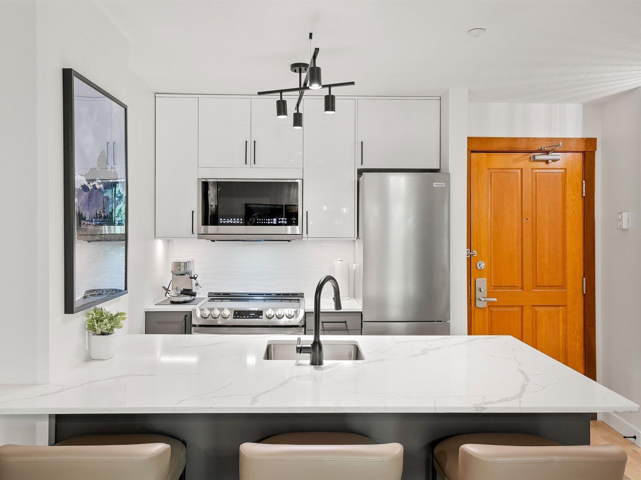 A modern kitchen with white cabinets, a stainless steel refrigerator, a built-in oven, a coffee maker, and a marble island with seating.
