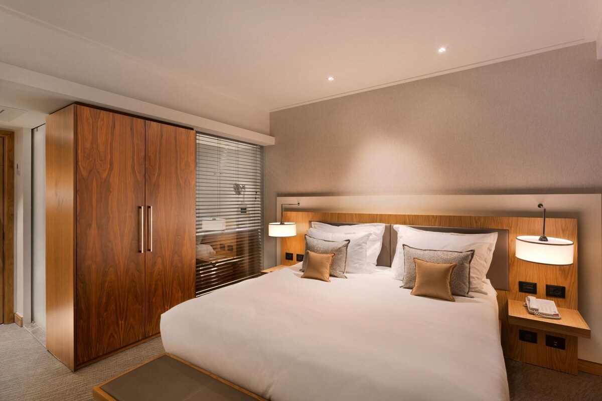 A modern hotel room with a large bed, wooden wardrobe, bedside lamps, and a view through the window blinds.