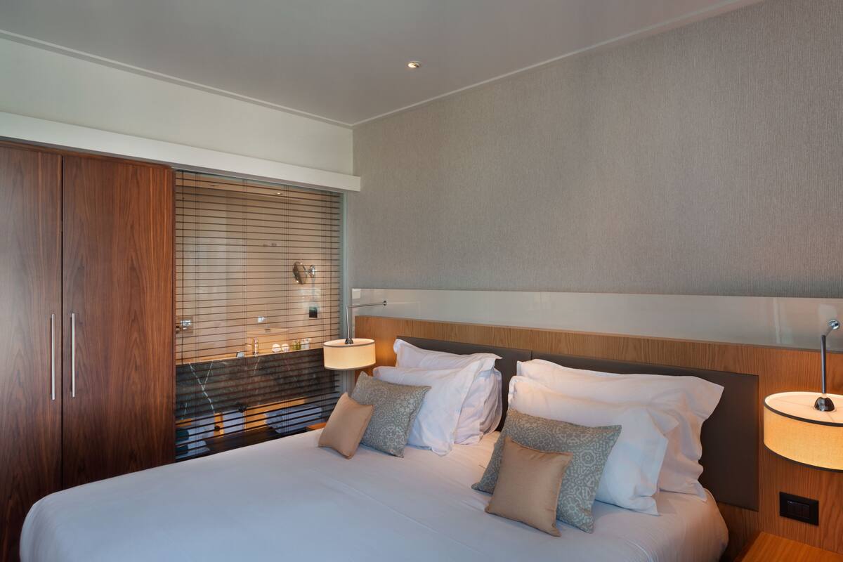 A modern hotel room with a large bed, wooden wardrobe, and a window with blinds.