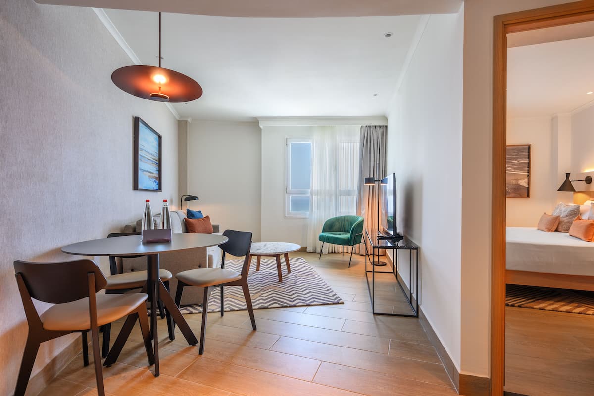A modern hotel room with a dining area, a green armchair, a sofa, and a bed.