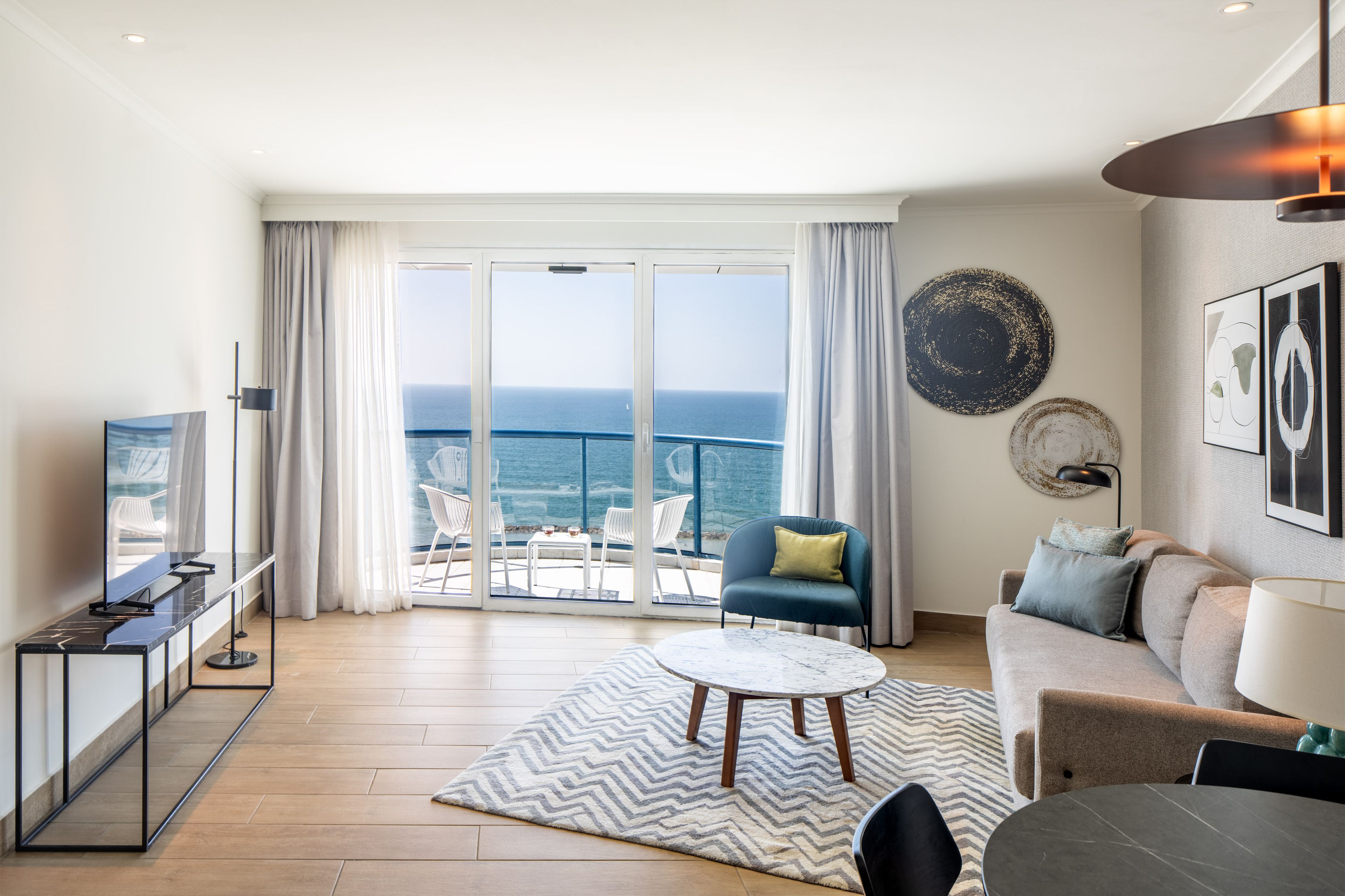 A modern living room with a large window, a flat-screen TV, a sofa, a coffee table, and a view of the sea.