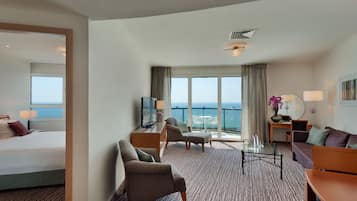 Une chambre d’hôtel comprenant un lit, deux fauteuils, un canapé, une table basse et un téléviseur. La chambre dispose de grandes fenêtres donnant sur la mer.