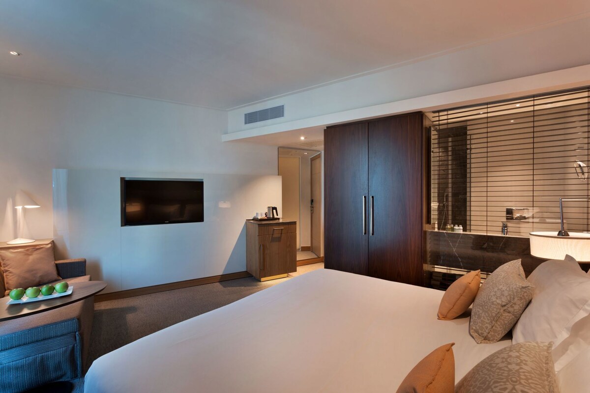 A hotel room with a bed, a TV, a desk with a lamp, a wardrobe, and a bathroom with a mirror.