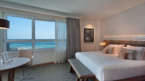 A hotel room with a large bed, a desk with a chair, a bench, a view of the ocean, and a framed picture on the wall. - Sea Tower by Isrotel Design (Tel Aviv)