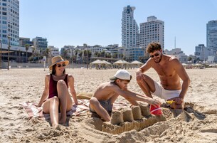 Escena de playa con gente construyendo castillos de arena, cielo despejado y edificios altos al fondo.