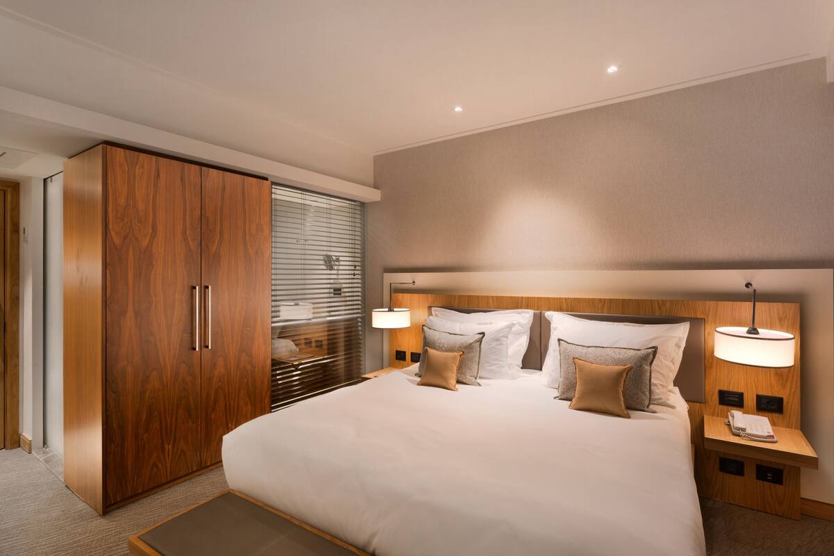 A modern hotel room with a large bed, wooden wardrobe, bedside lamps, and a view through the window blinds.