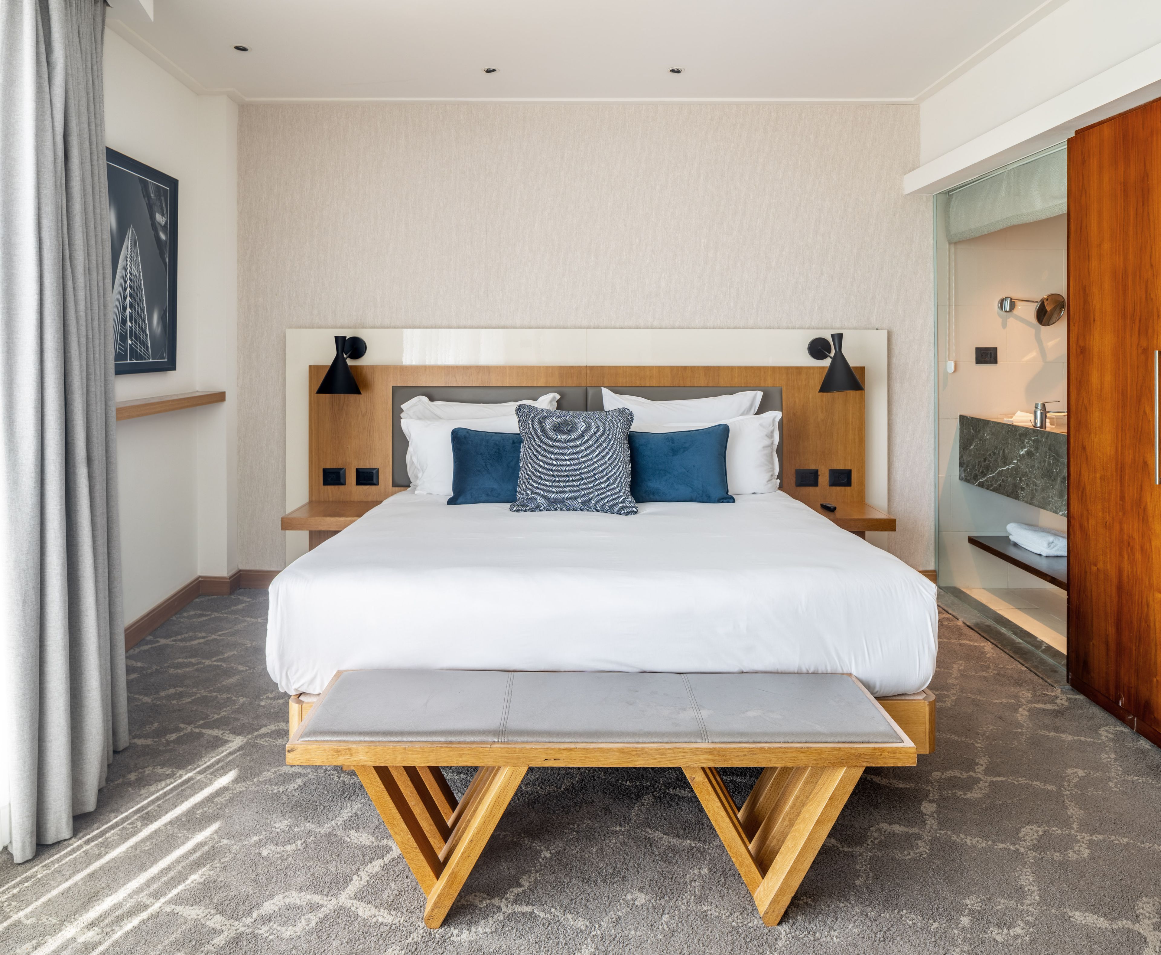 Une chambre d’hôtel avec un grand lit, des tables de chevet, un banc, une salle de bain avec un miroir et une vue sur un immeuble.