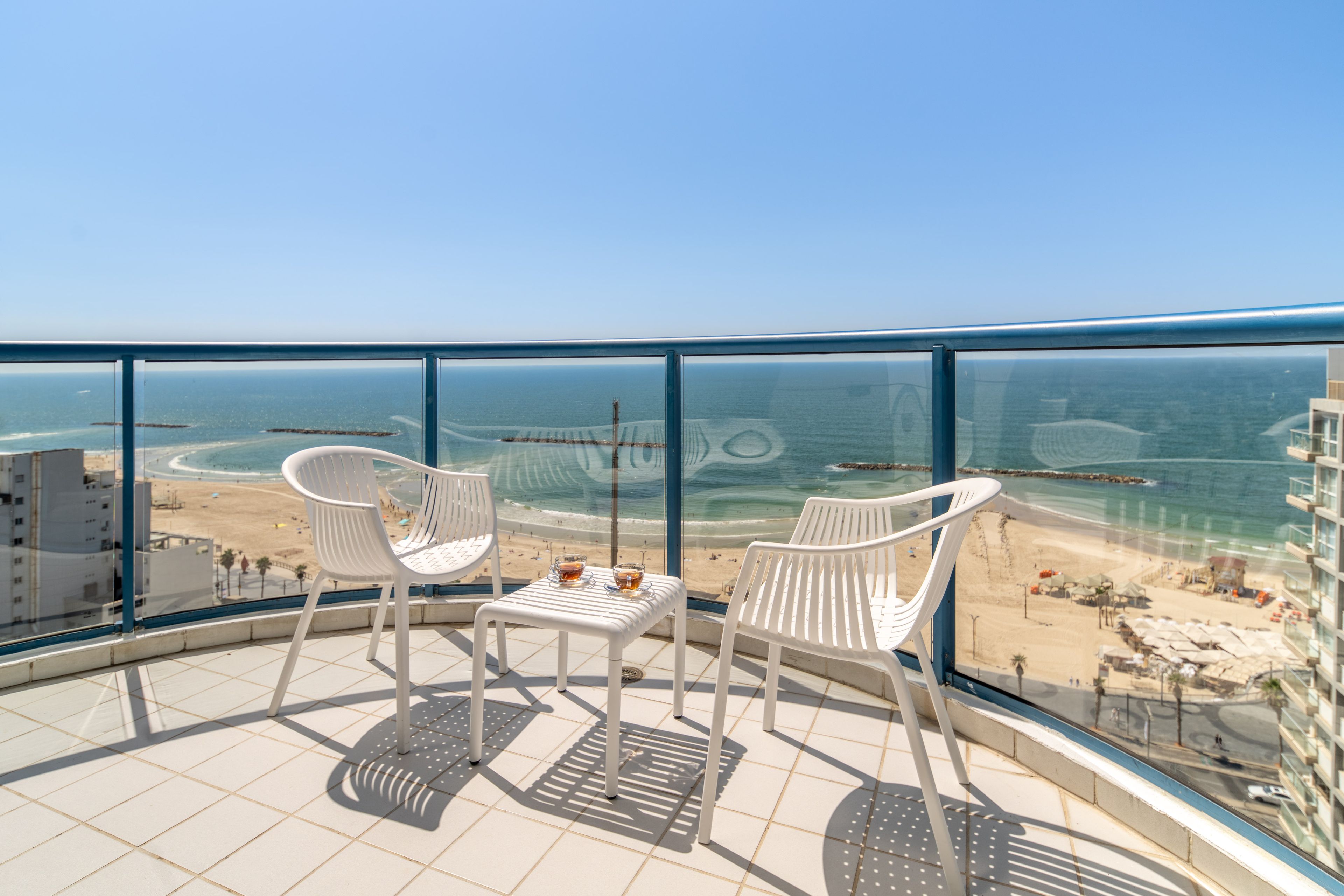 A balcony with two white chairs and a small table, overlooking a beach and ocean.