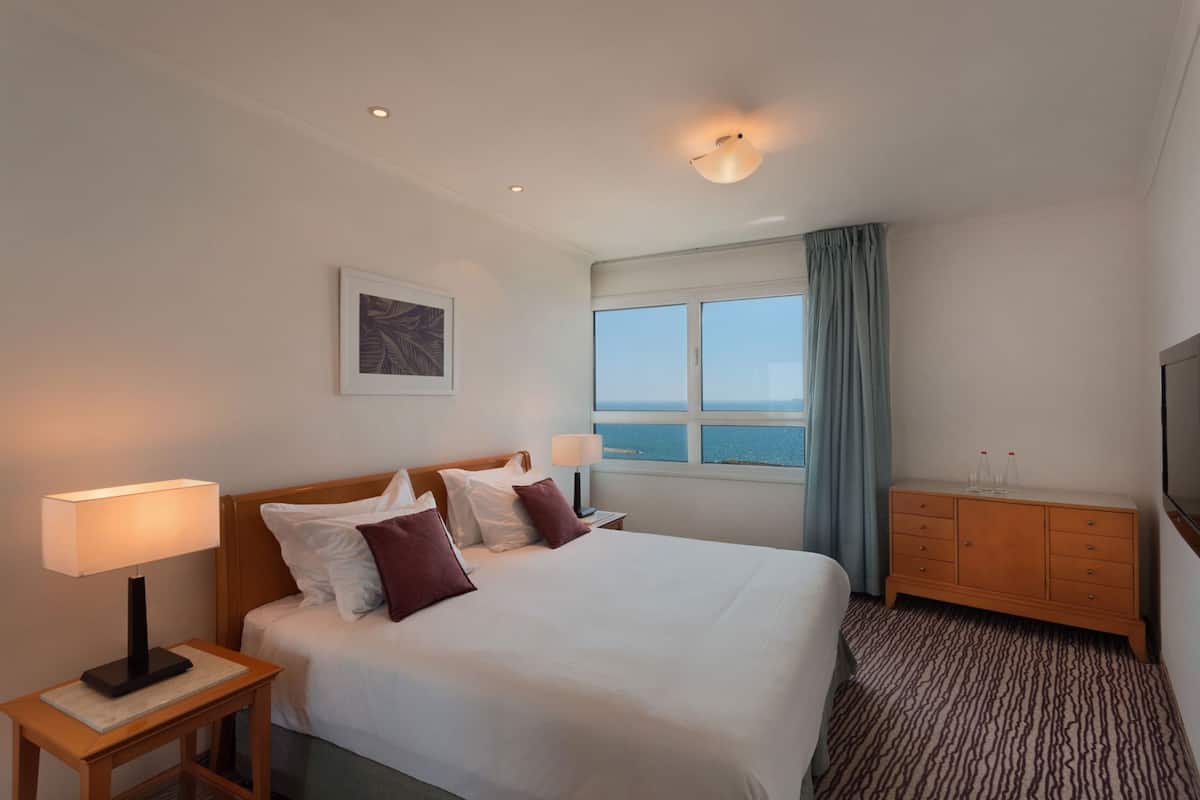 A hotel room with a bed, two bedside lamps, a wooden nightstand, a dresser, a television, a window with a view of the sea, and a ceiling light.