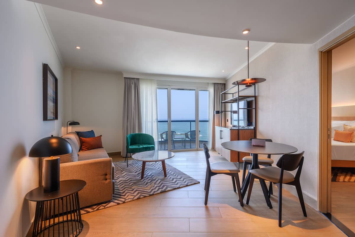 A modern hotel room with a sofa, a dining table with chairs, a small coffee table, a bookshelf, and a view of the sea through large windows.