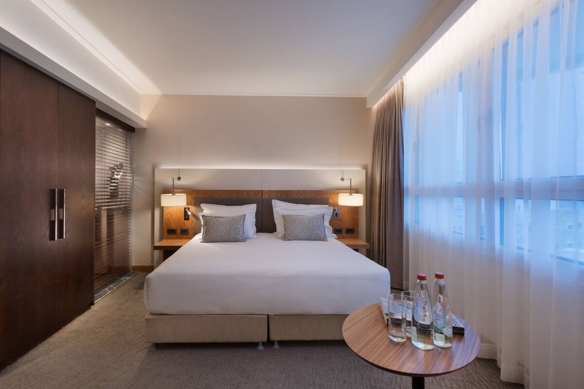 A hotel room with a large bed, two bedside tables with lamps, a wooden wardrobe, and a round table with bottles and glasses.