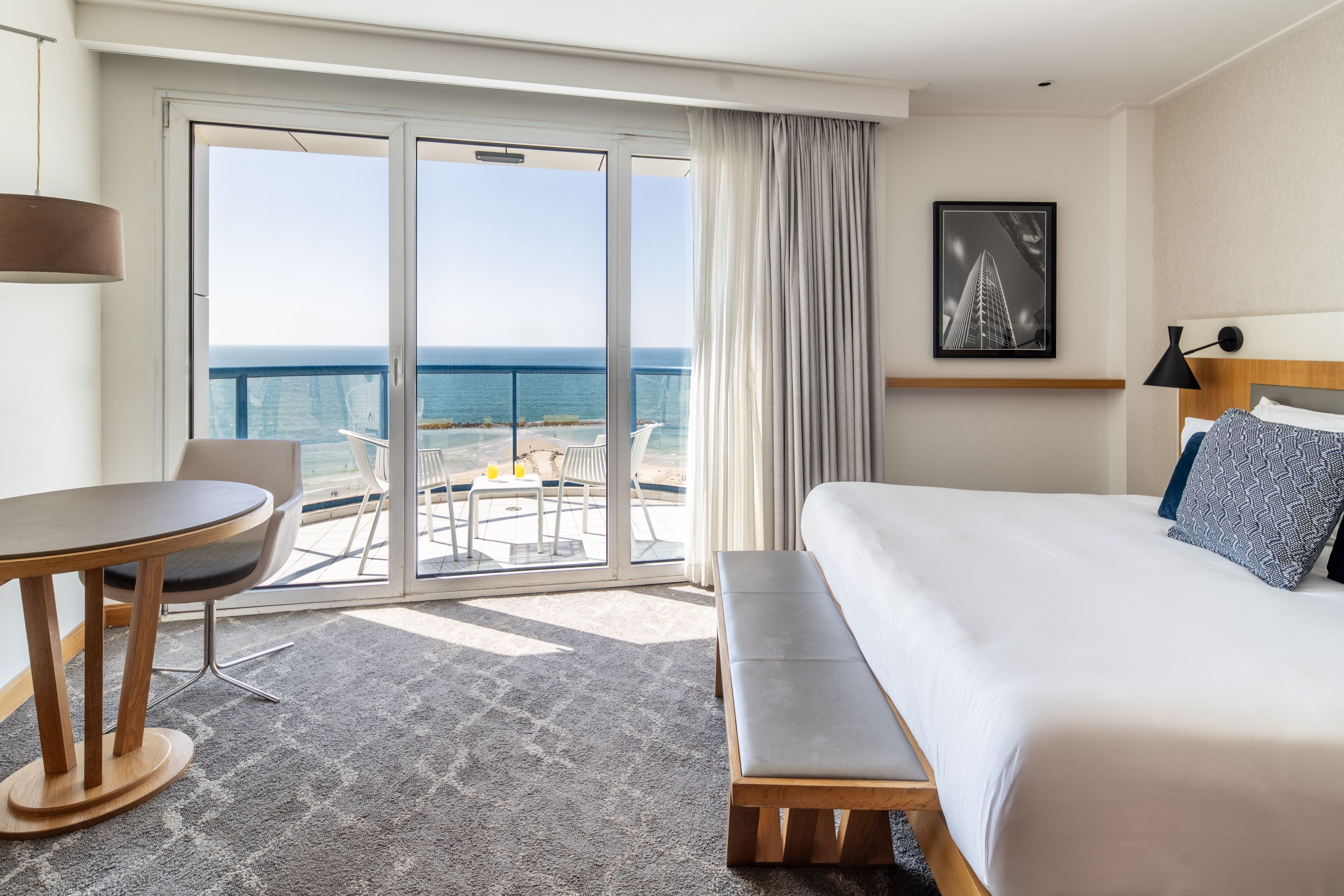 A hotel room with a bed, a desk, a chair, a bench, a table with two chairs, a large window with a view of the sea, and a framed picture on the wall.