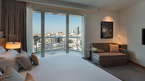 A hotel room with a large bed, a sofa, a balcony with a city view, and a framed picture on the wall. - Sea Tower by Isrotel Design (Tel Aviv)