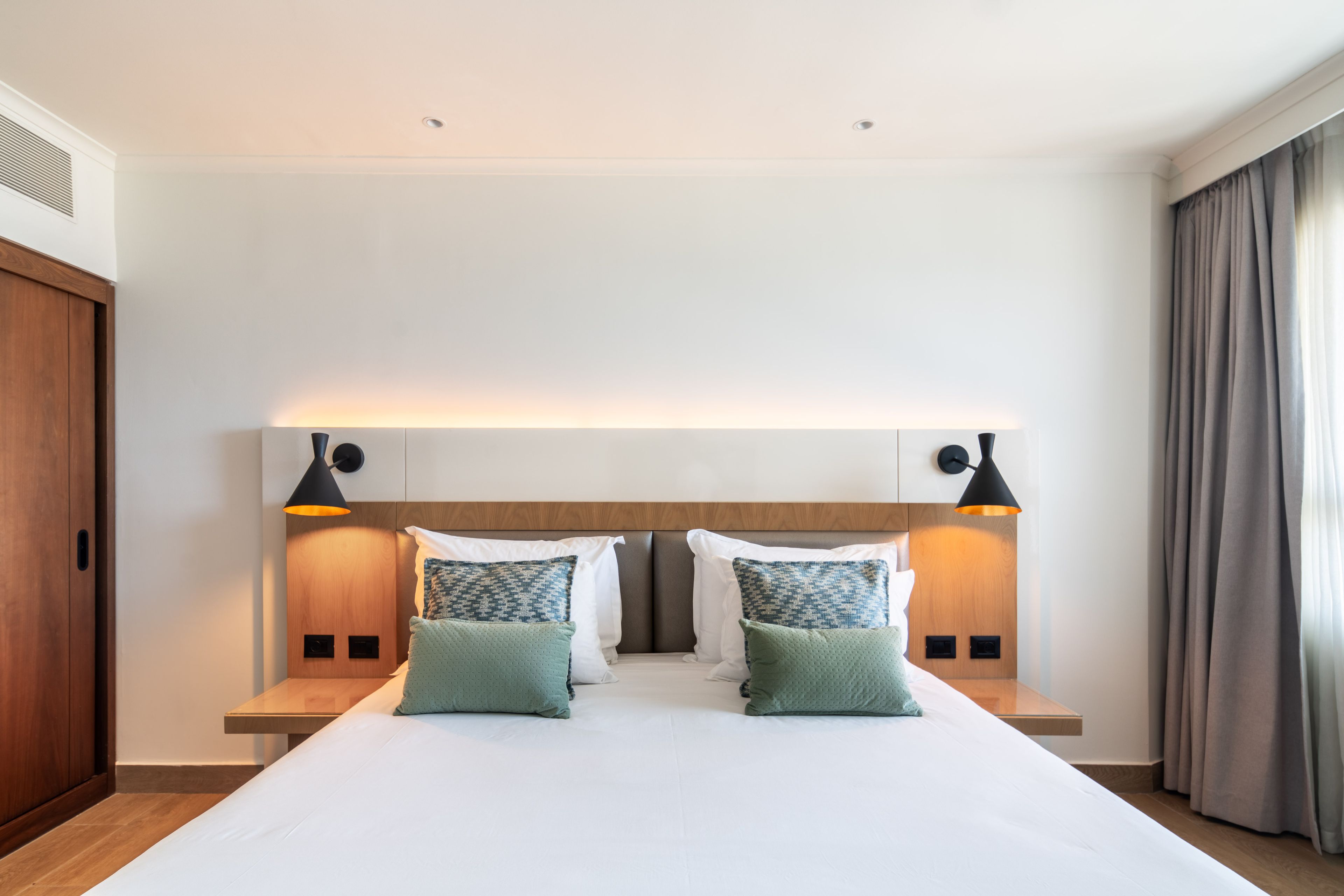 Habitación de hotel con una cama grande, dos mesitas de noche, un cabecero con iluminación integrada y vistas al exterior a través de una ventana con cortinas.
