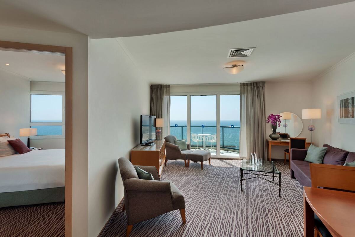 A hotel room with a bed, a TV, a desk with a chair, a sofa, and a balcony with a view of the sea.