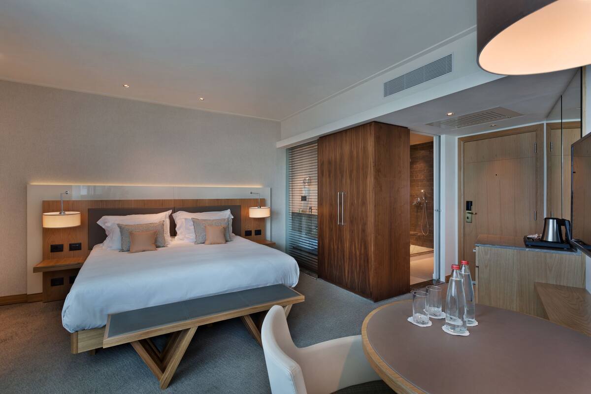 A modern hotel room with a large bed, bedside tables, a desk, and a dining area.