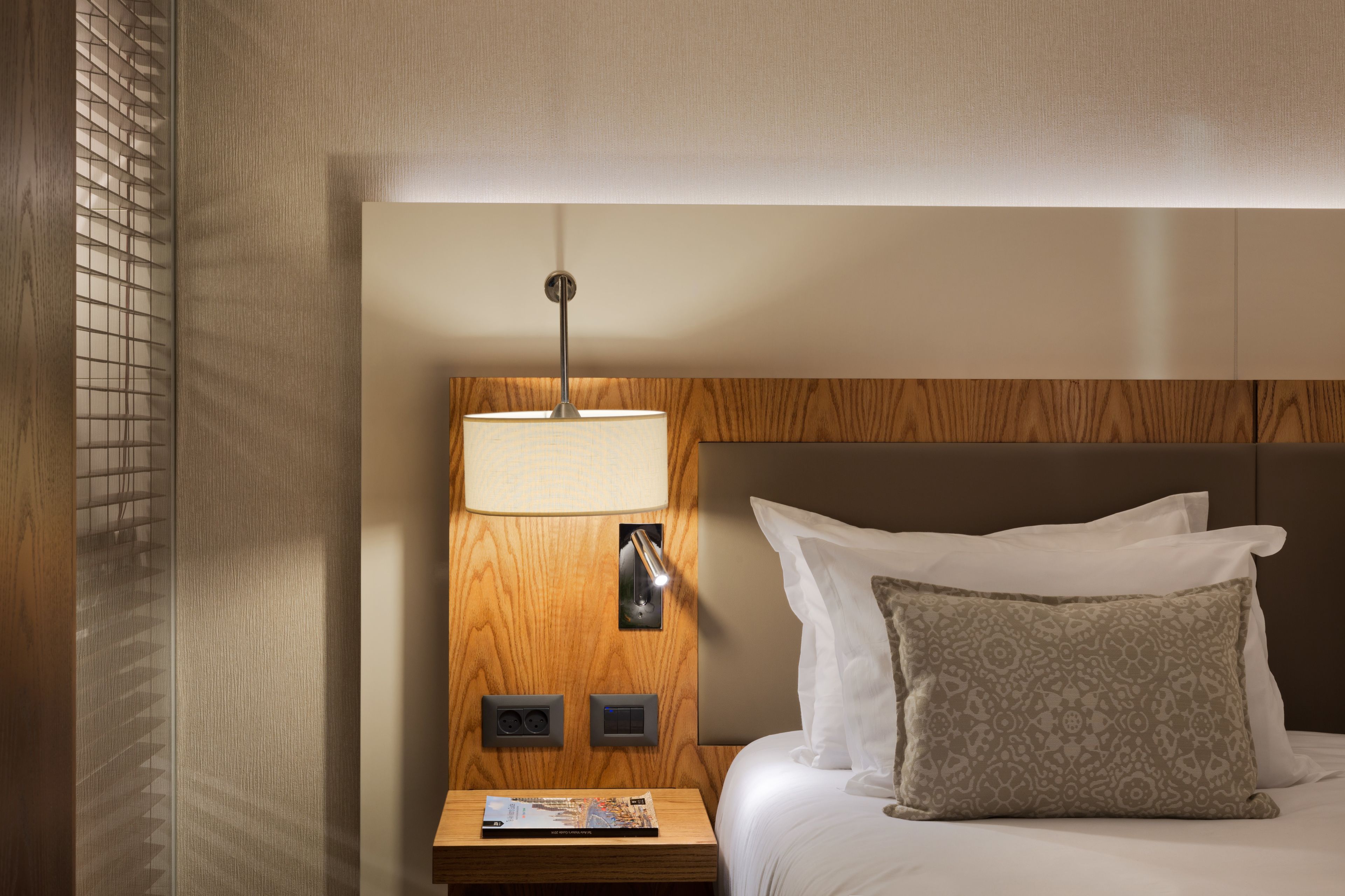 A hotel room with a wooden headboard, a bedside lamp, a nightstand with a magazine, and a patterned pillow.